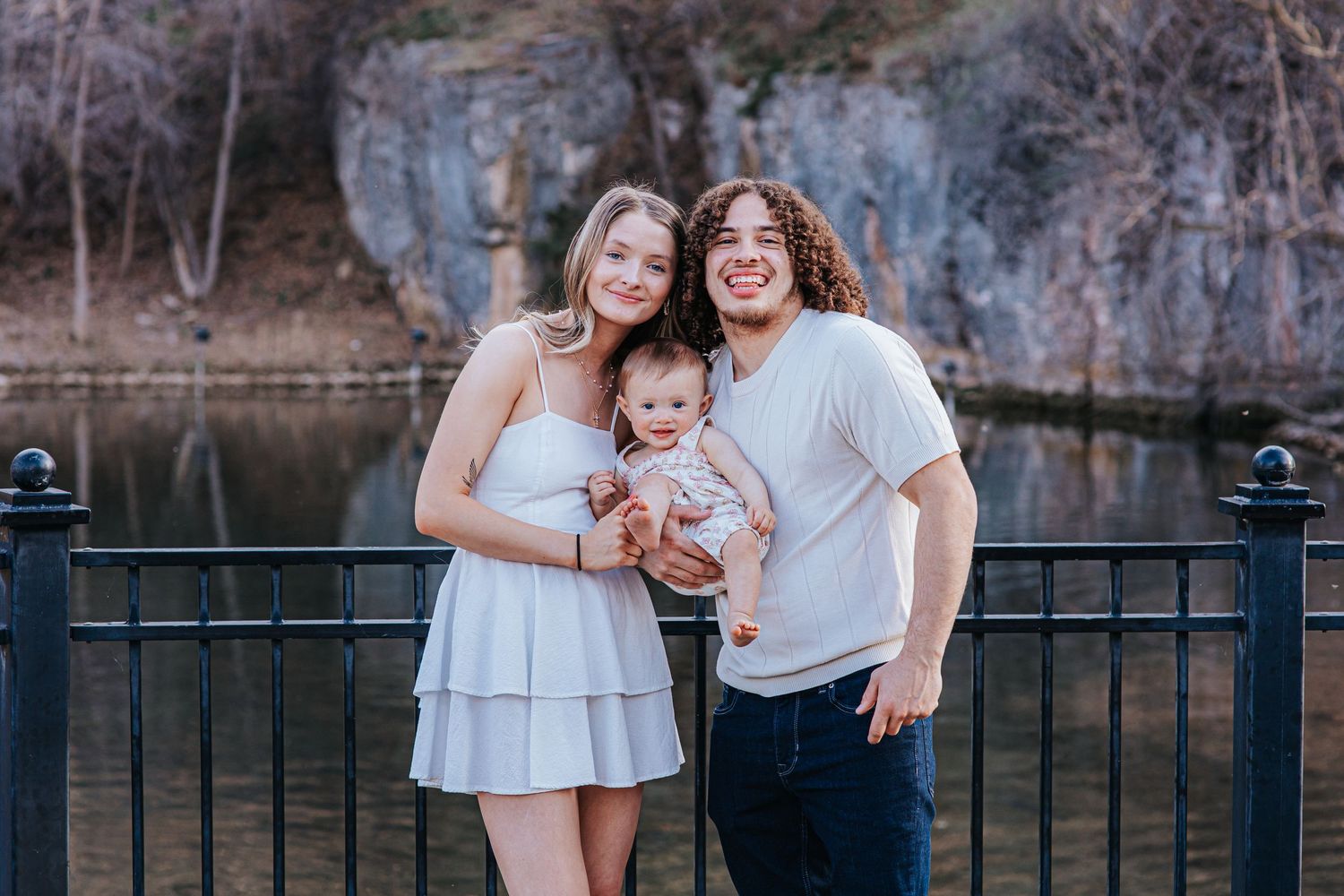 Young couple smiling outdoors by a railing, holding a baby, with a scenic river and rocky cliffs in the background.