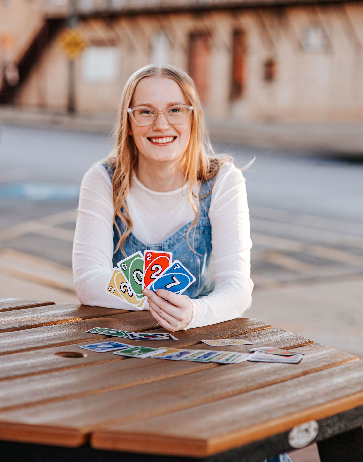 Smiling young woman with glasses holding colorful UNO cards at an outdoor wooden table.