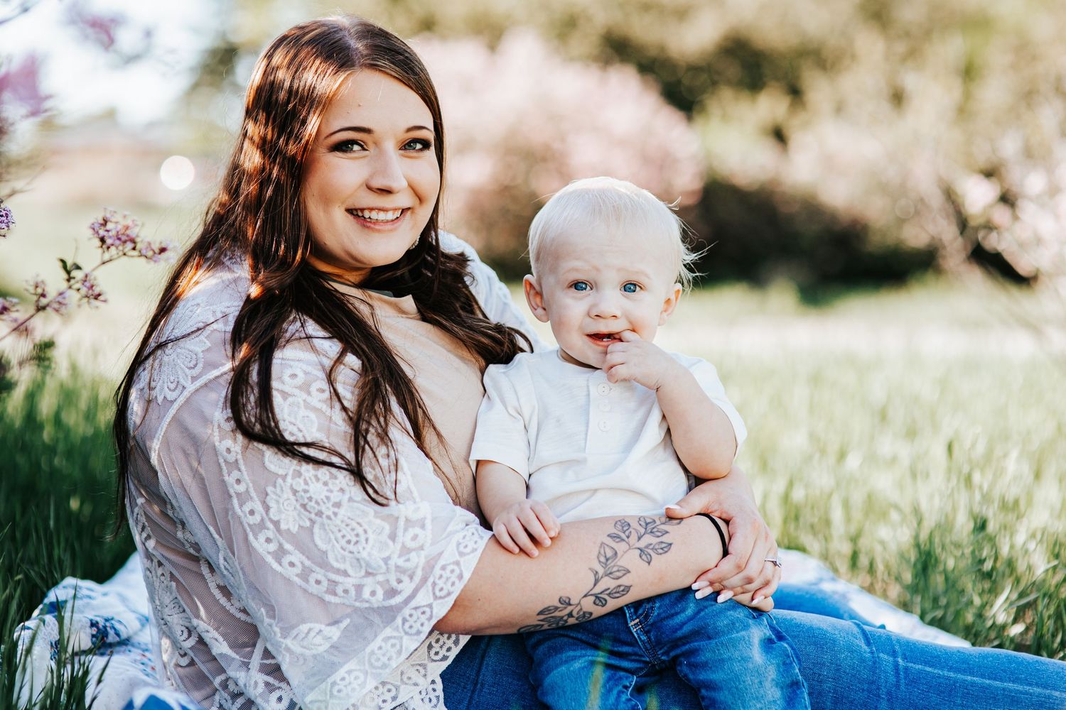 A parent and baby sit together in a sunny meadow wearing white outfits during a springtime outdoor photo session.
