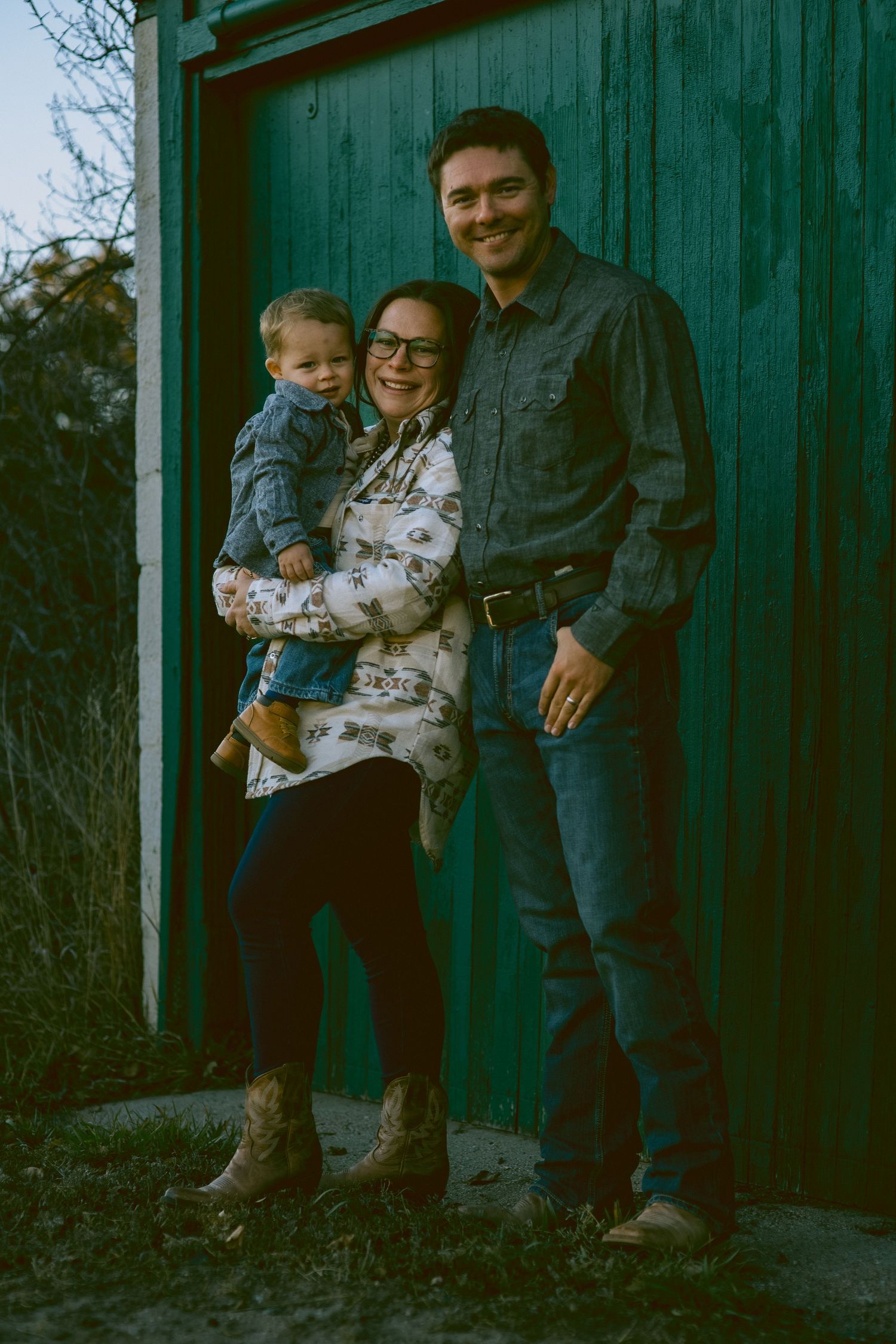 A smiling family of three poses outdoors by a teal barn, mother holding toddler, father beside her, wearing casual western attire.