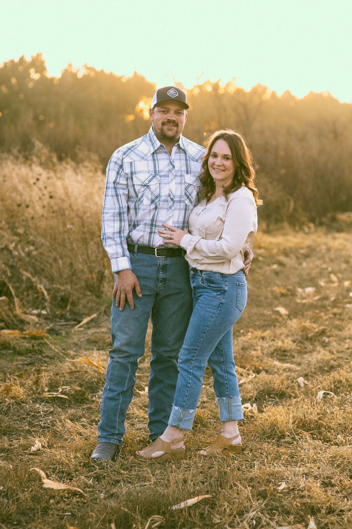 A couple in casual western wear poses together at sunset in a rustic outdoor setting in a corn field in Nebraska
