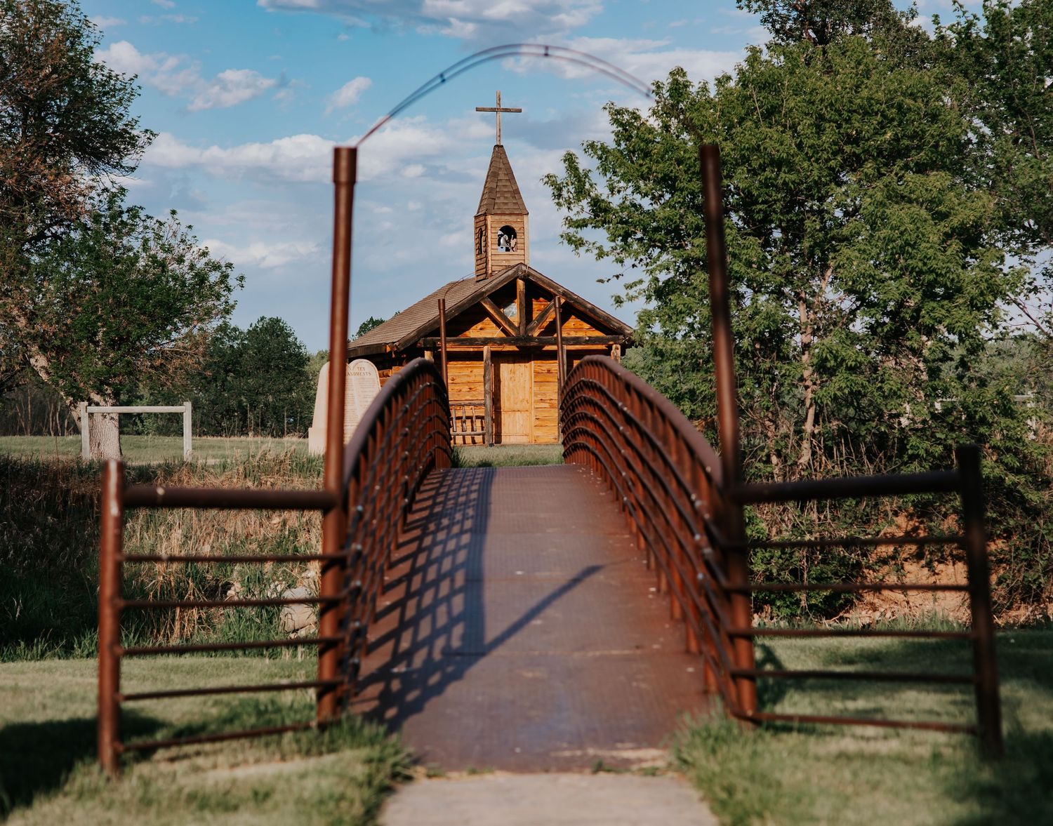 A rustic wooden bridge leads to a charming log chapel with a cross steeple, surrounded by lush green trees under a blue sky.