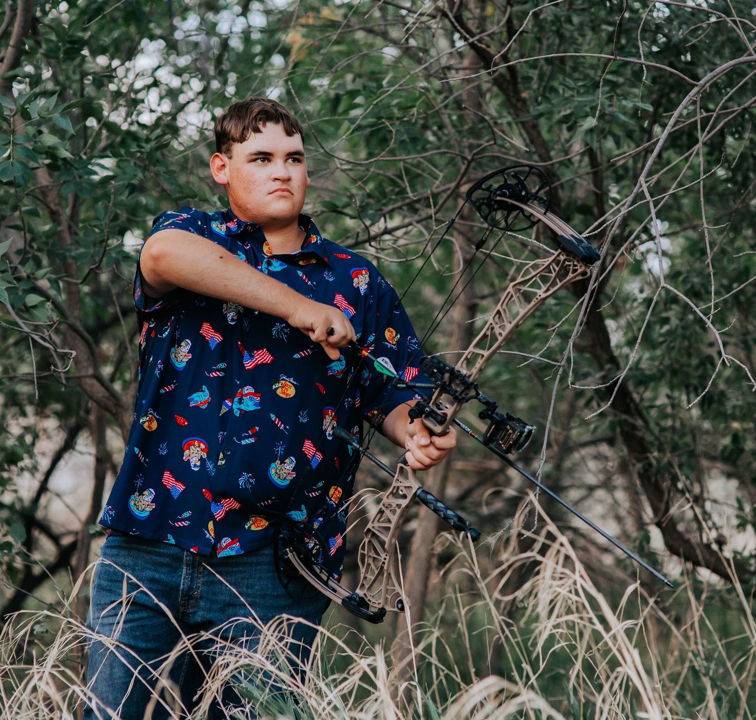 Young man in colorful shirt drawing a compound bow outdoors among tall grass and trees.