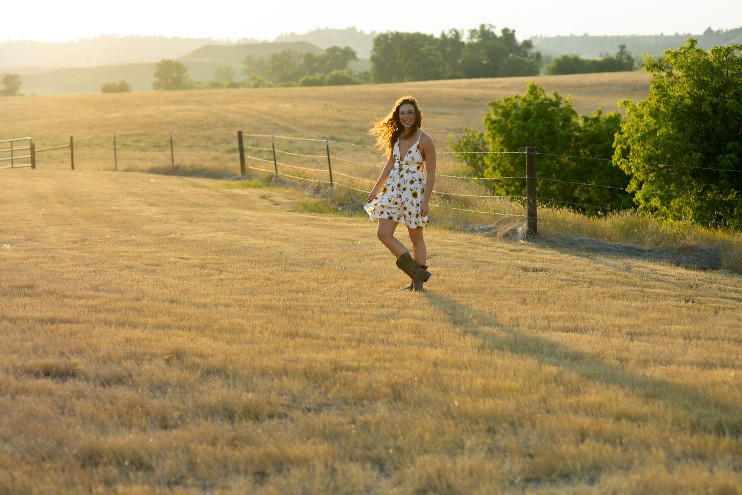 Young woman in floral dress running through golden sunlit meadow with fence and trees in background.