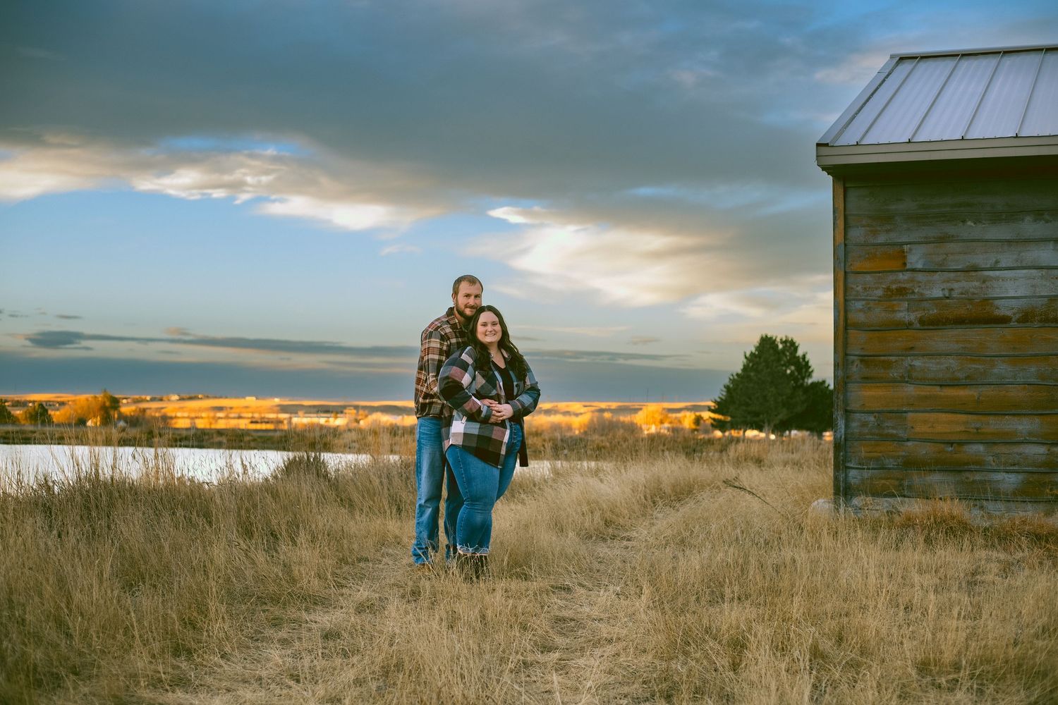 Scenic panoramic view of engaged couple standing in golden prairie grass near rustic wooden buildings at sunset in Western Nebraska.