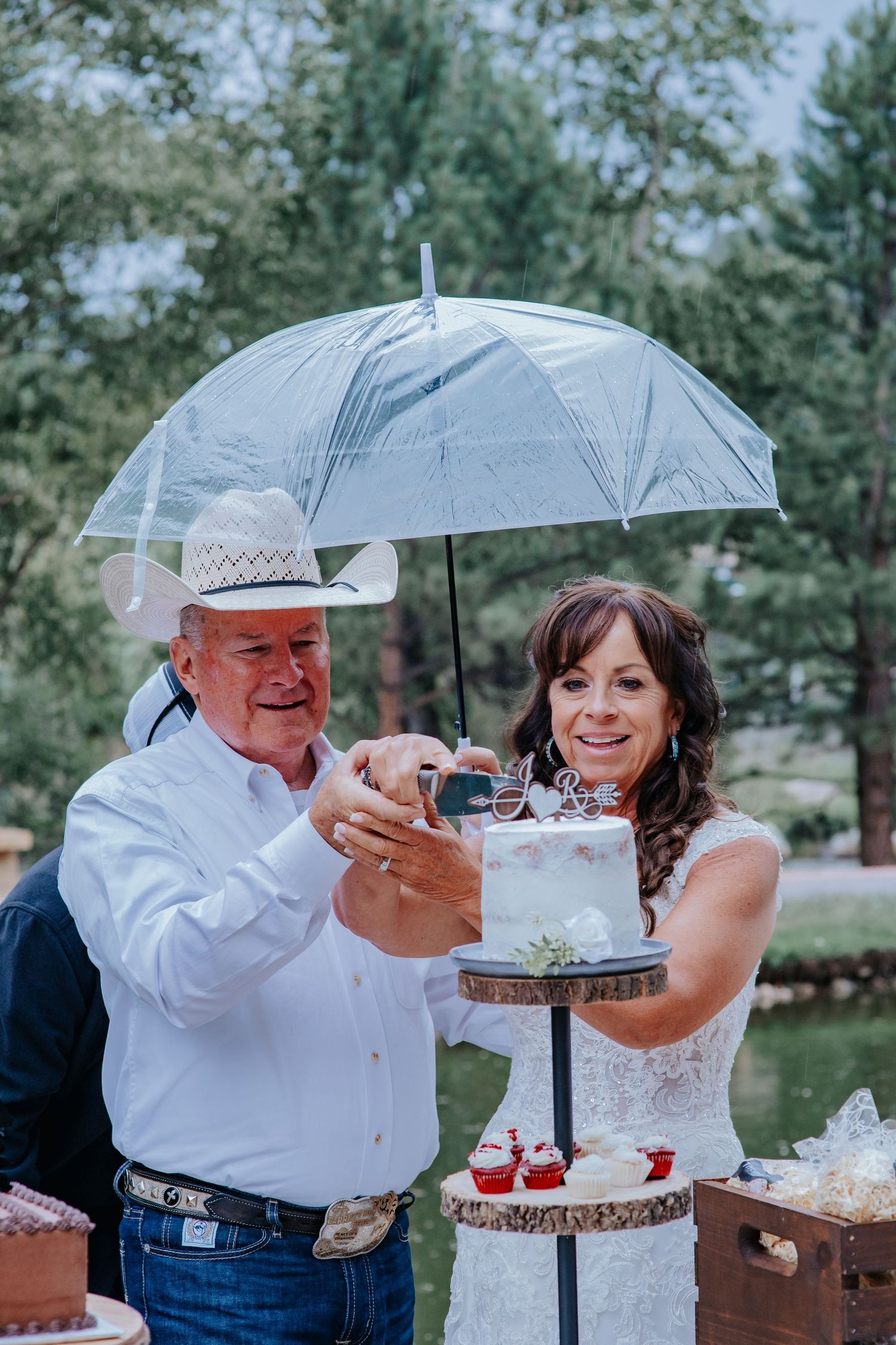 A couple cuts their wedding cake together under a clear umbrella at an outdoor rustic-themed celebration in Estes Park Colorado