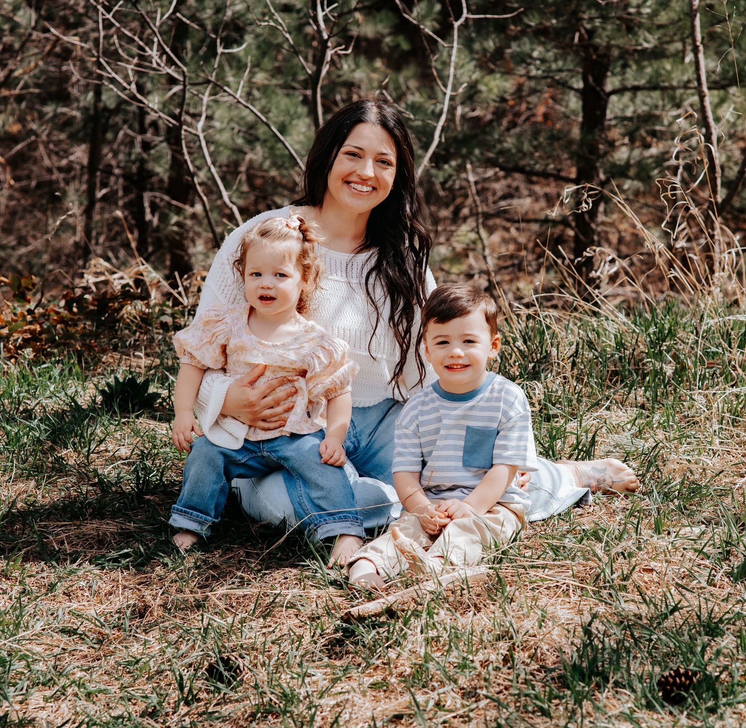 A smiling woman sits outdoors with two young children on grass surrounded by trees in a warm family portrait.