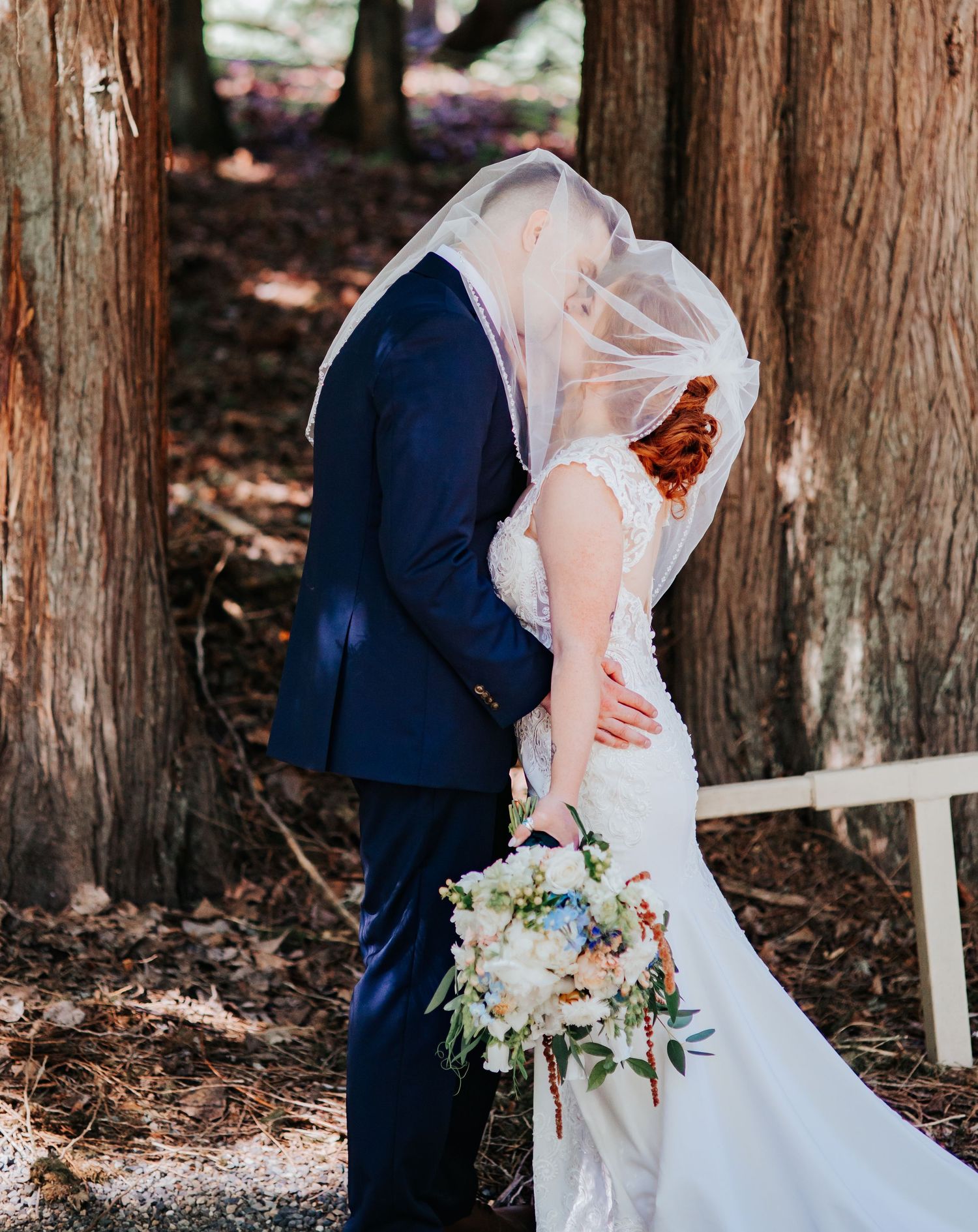 Newlyweds share an intimate moment between redwood trees while holding a pastel floral bouquet.