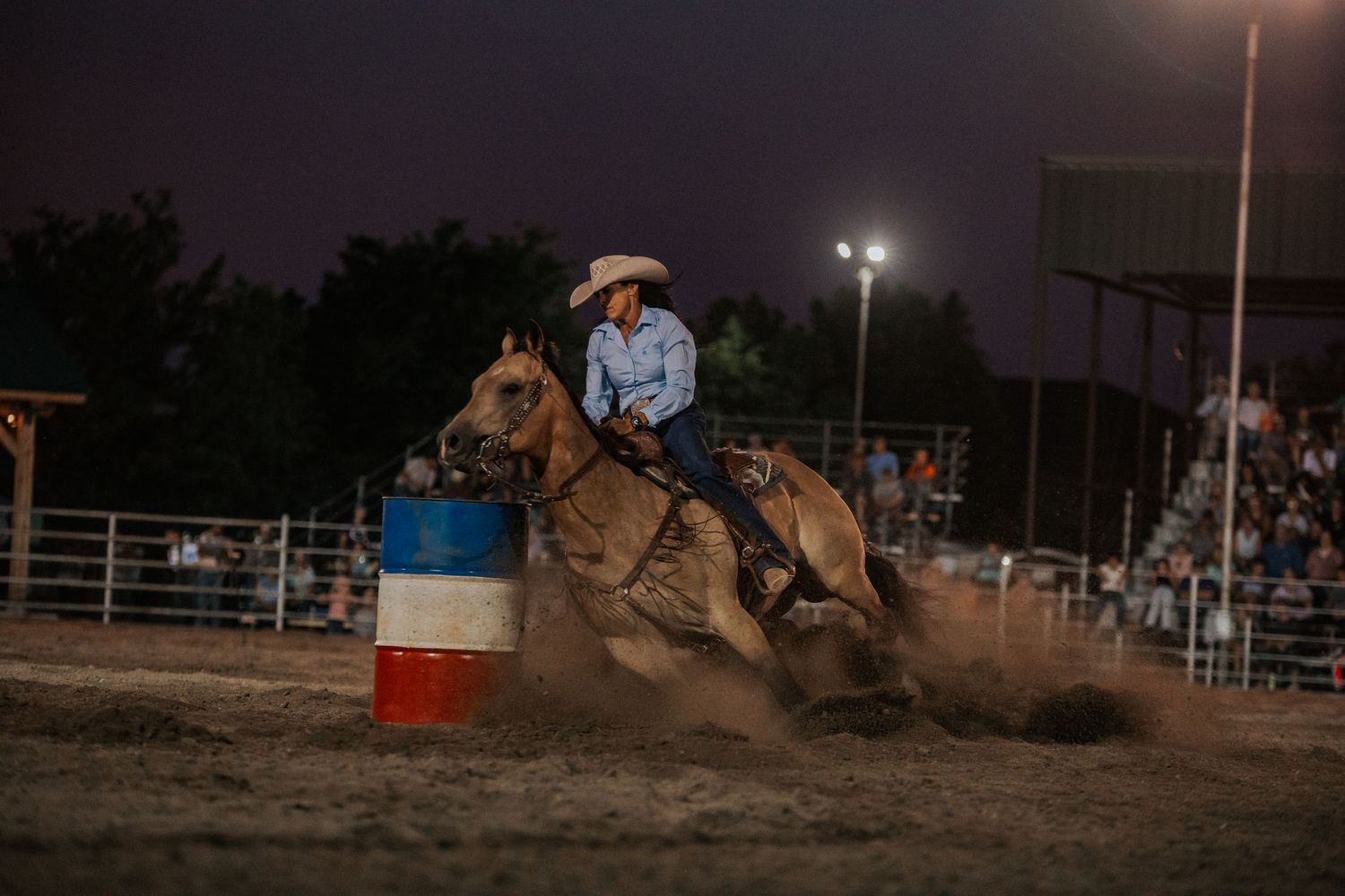 Dramatic nighttime rodeo scene showing barrel racing action in a dusty arena.
