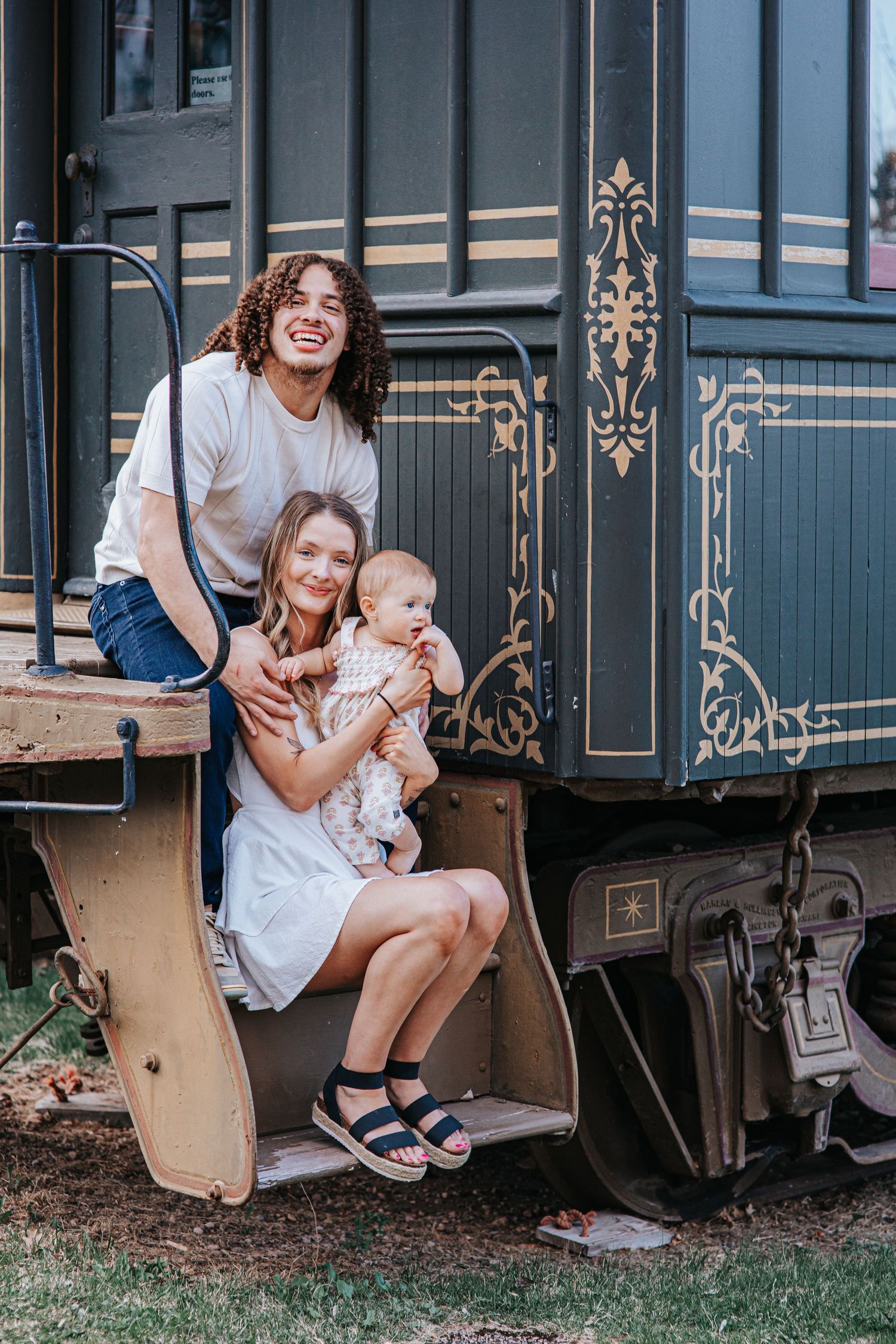 Young family with baby posing on vintage train car steps, smiling outdoors on a sunny day.
