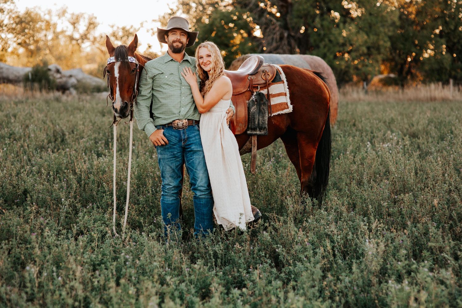 A couple in western attire stands with their brown horse in a sunlit meadow during golden hour in Nebraska