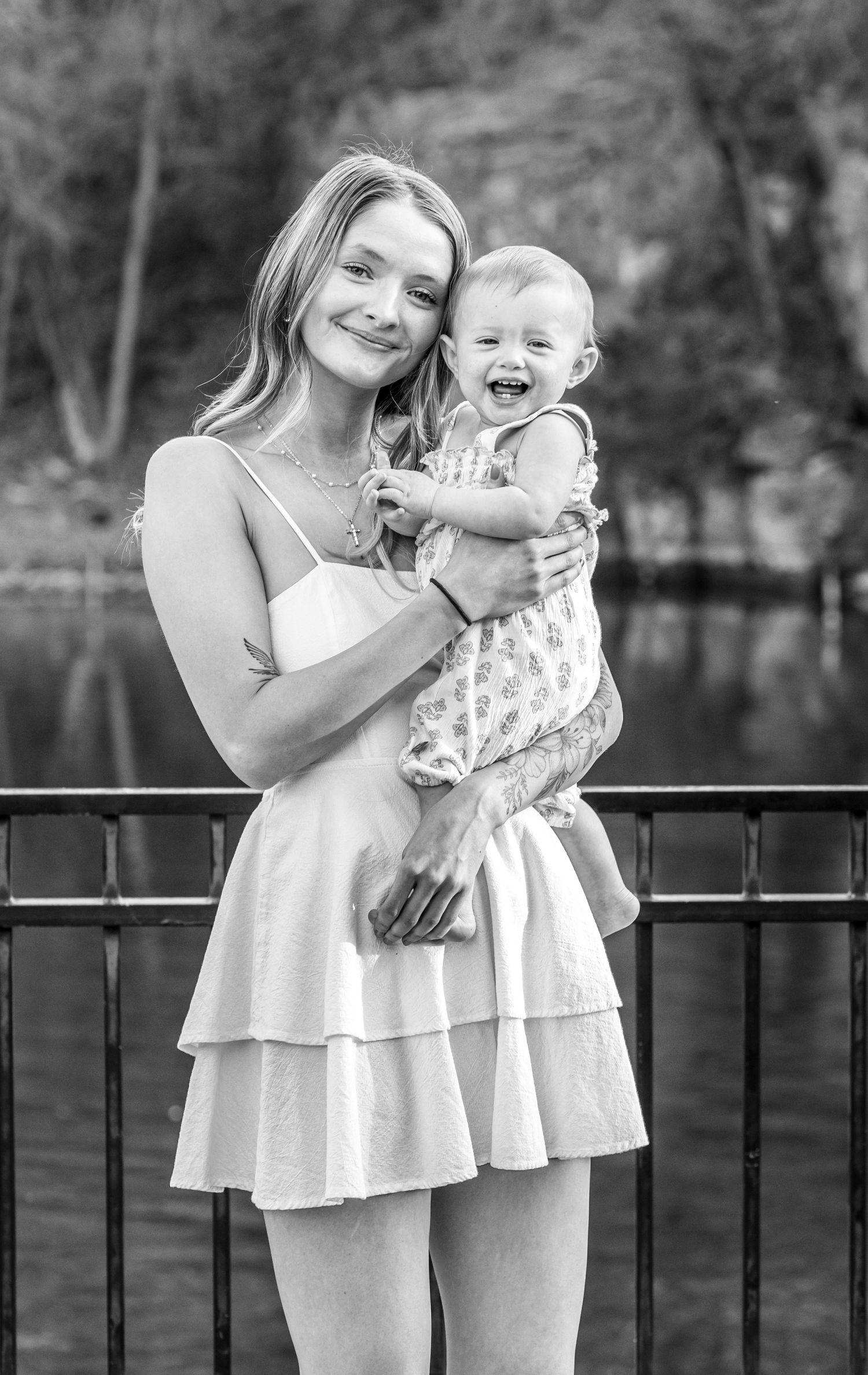 Black and white photo of a young woman in a white dress holding a laughing baby near a railing outdoors.