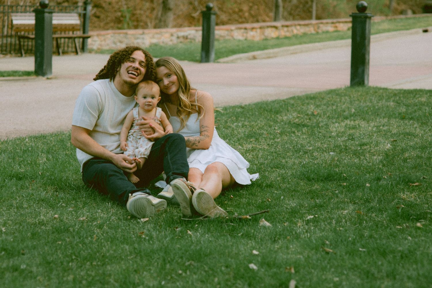 Happy family sitting on green grass in a park, parents smiling with baby between them on a sunny day.
