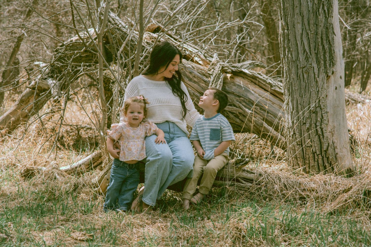 A mother sits with her two young children against a rustic fallen tree in a natural woodland setting.