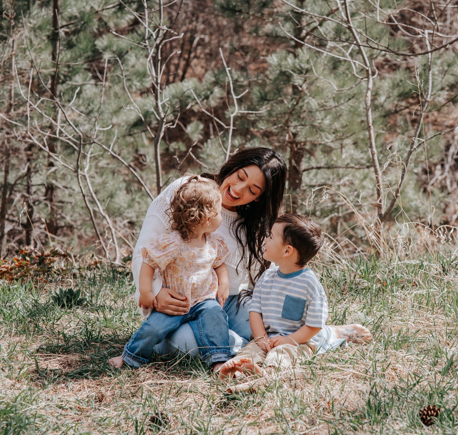 A mother sits on grass with her two young children, smiling warmly in a sunlit woodland setting.