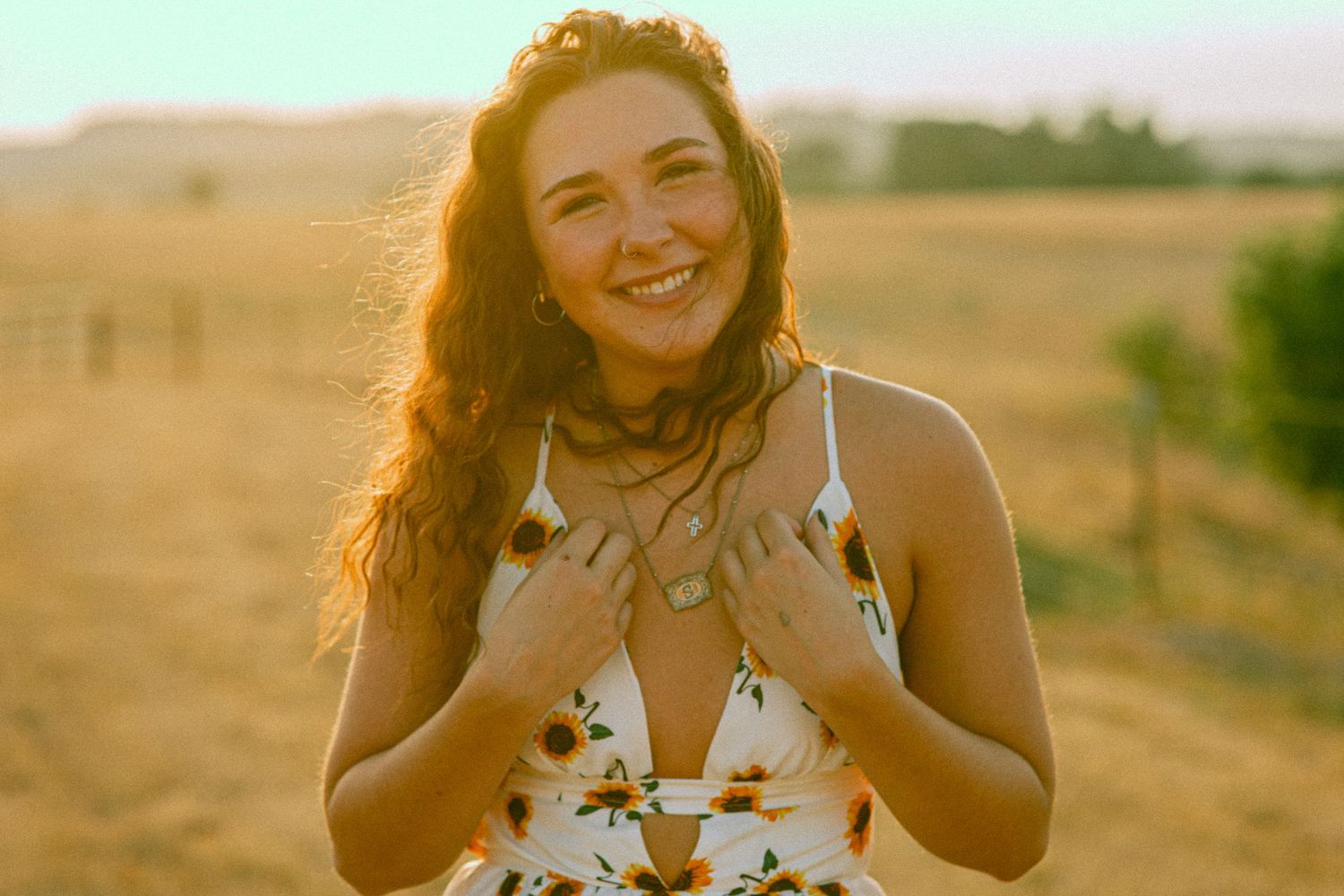 Young woman in floral sundress smiling joyfully in golden sunset light in an open field.