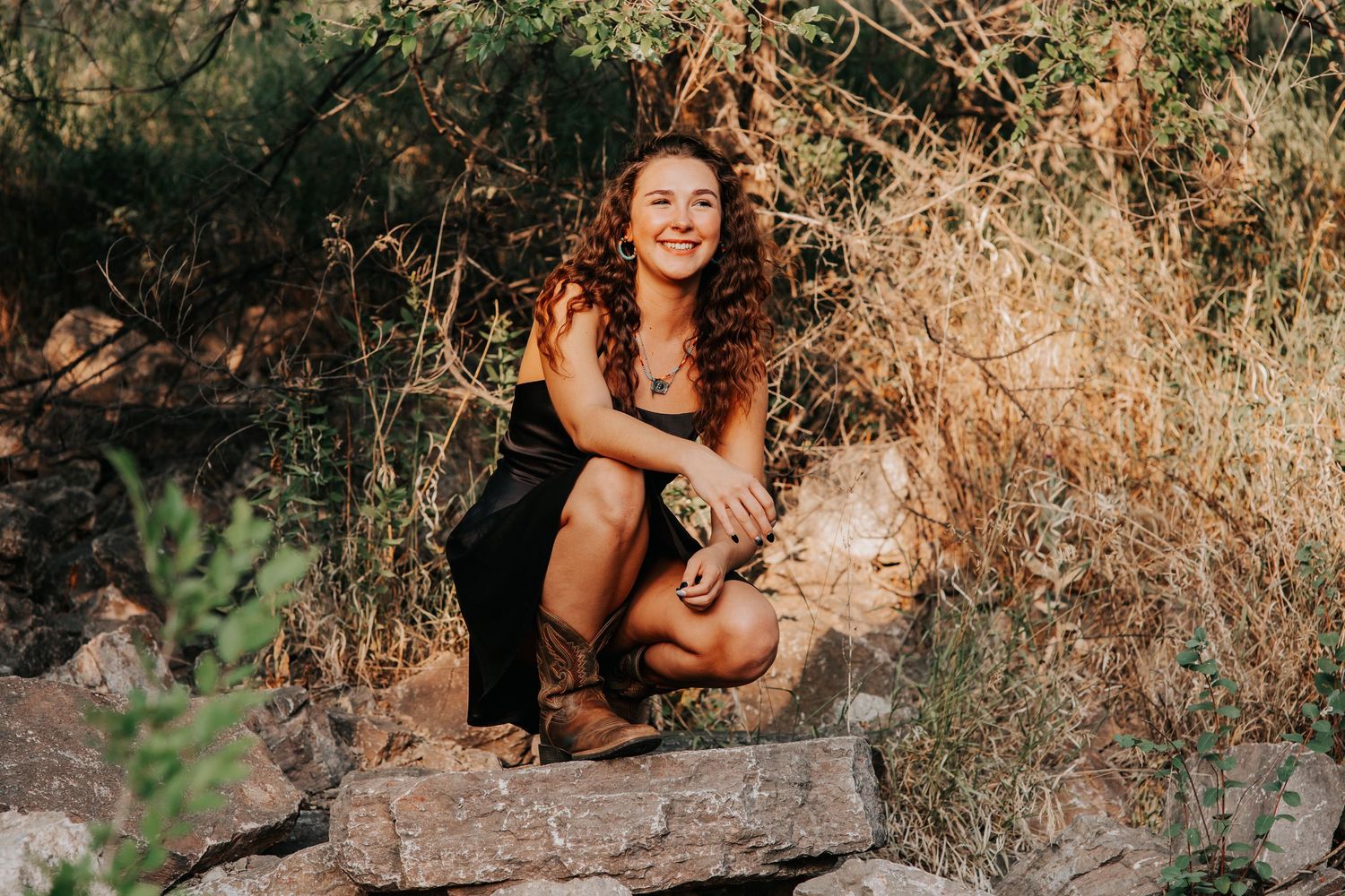 A young woman with curly hair smiles while crouching on rocks surrounded by dry brush in a natural outdoor setting.