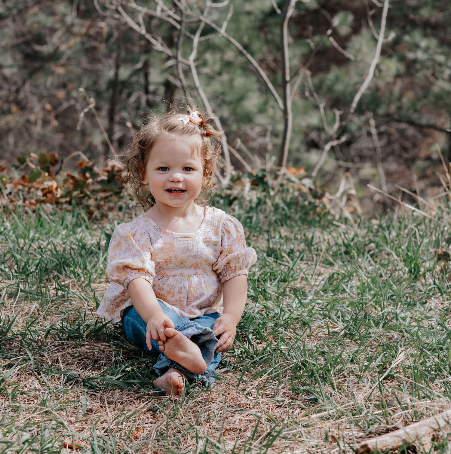 A smiling toddler girl sits cross-legged on grass in a floral top and jeans, with trees in the background.