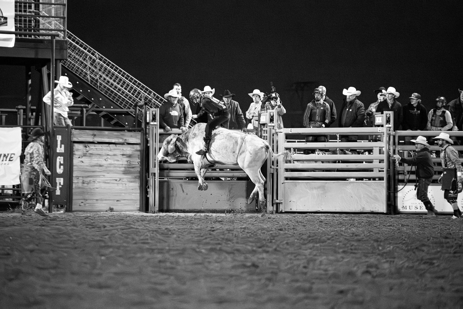 Black and white photo of spectators watching a rodeo event from behind wooden arena fencing.