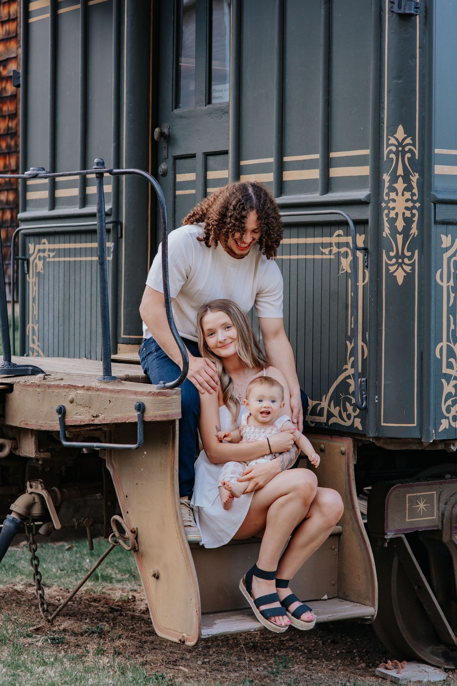 Young family with curly-haired dad, mom in white dress, and baby poses on vintage train car steps outdoors.