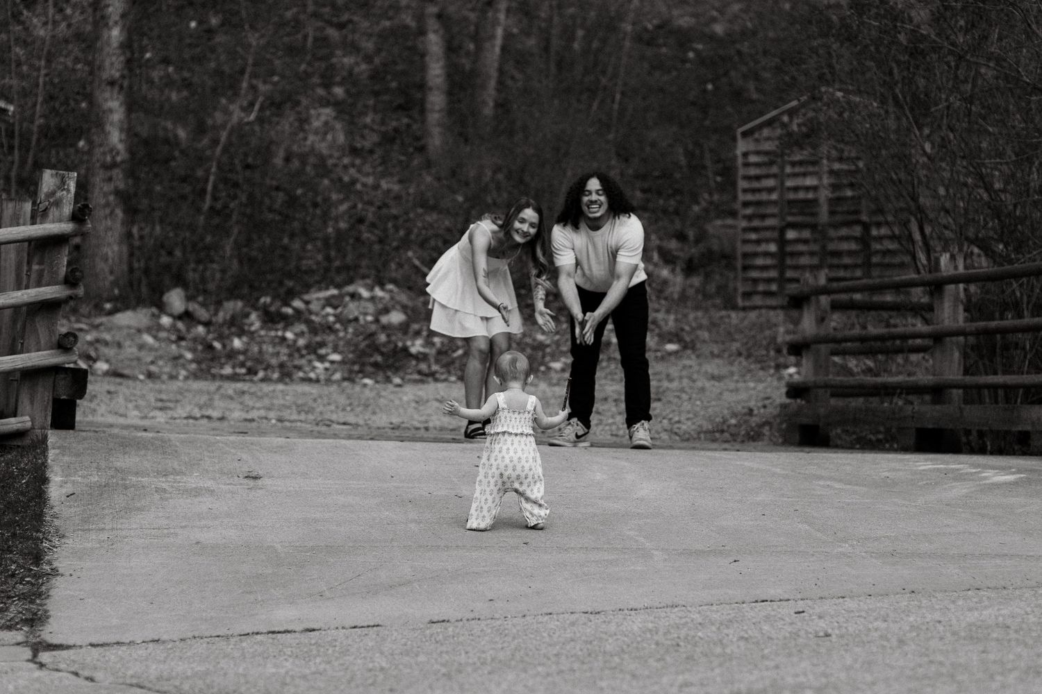Black and white photo of two women encouraging a toddler taking first steps on a paved path outdoors.
