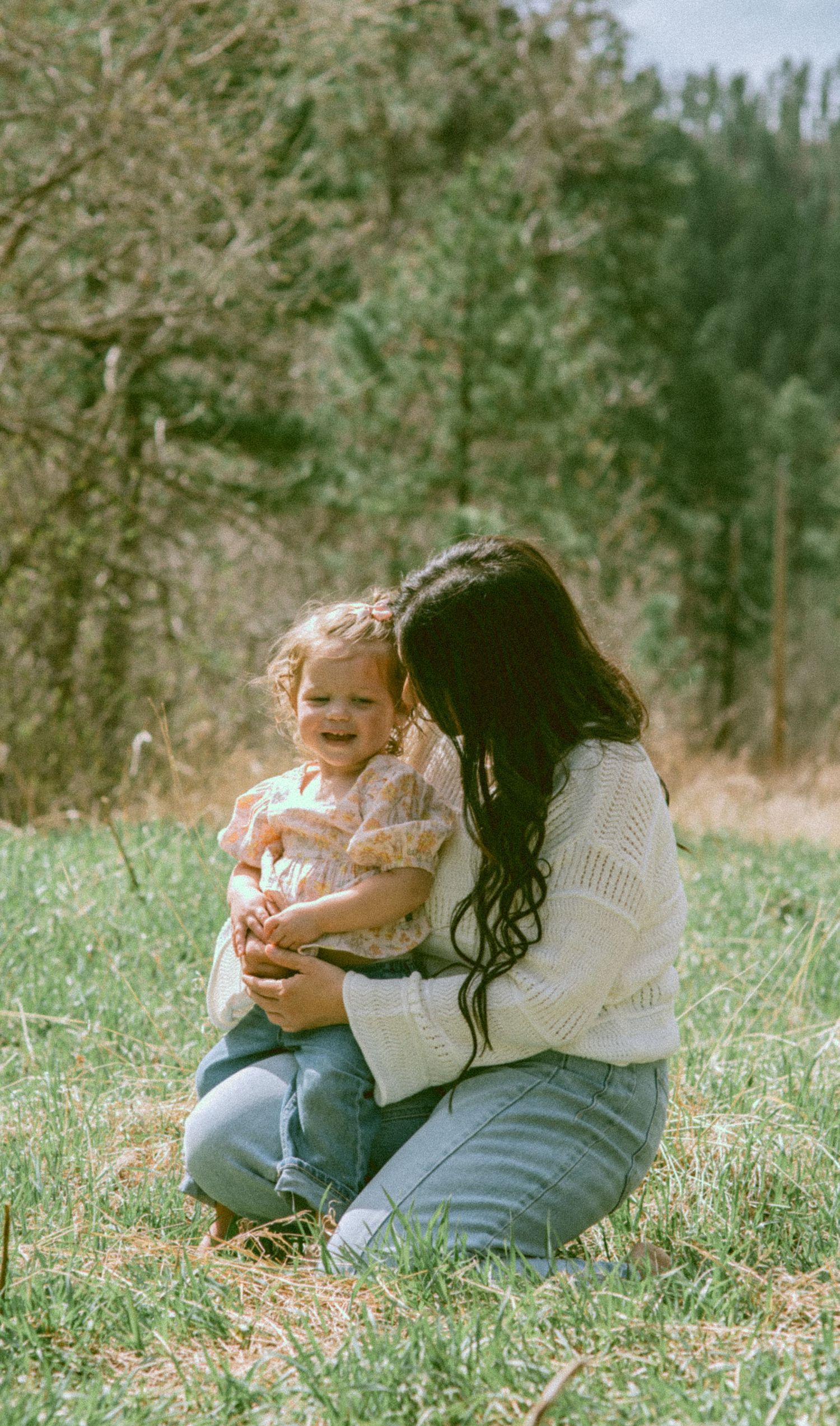 A mother with long dark hair sits on grass hugging her smiling toddler outdoors with trees in the background.