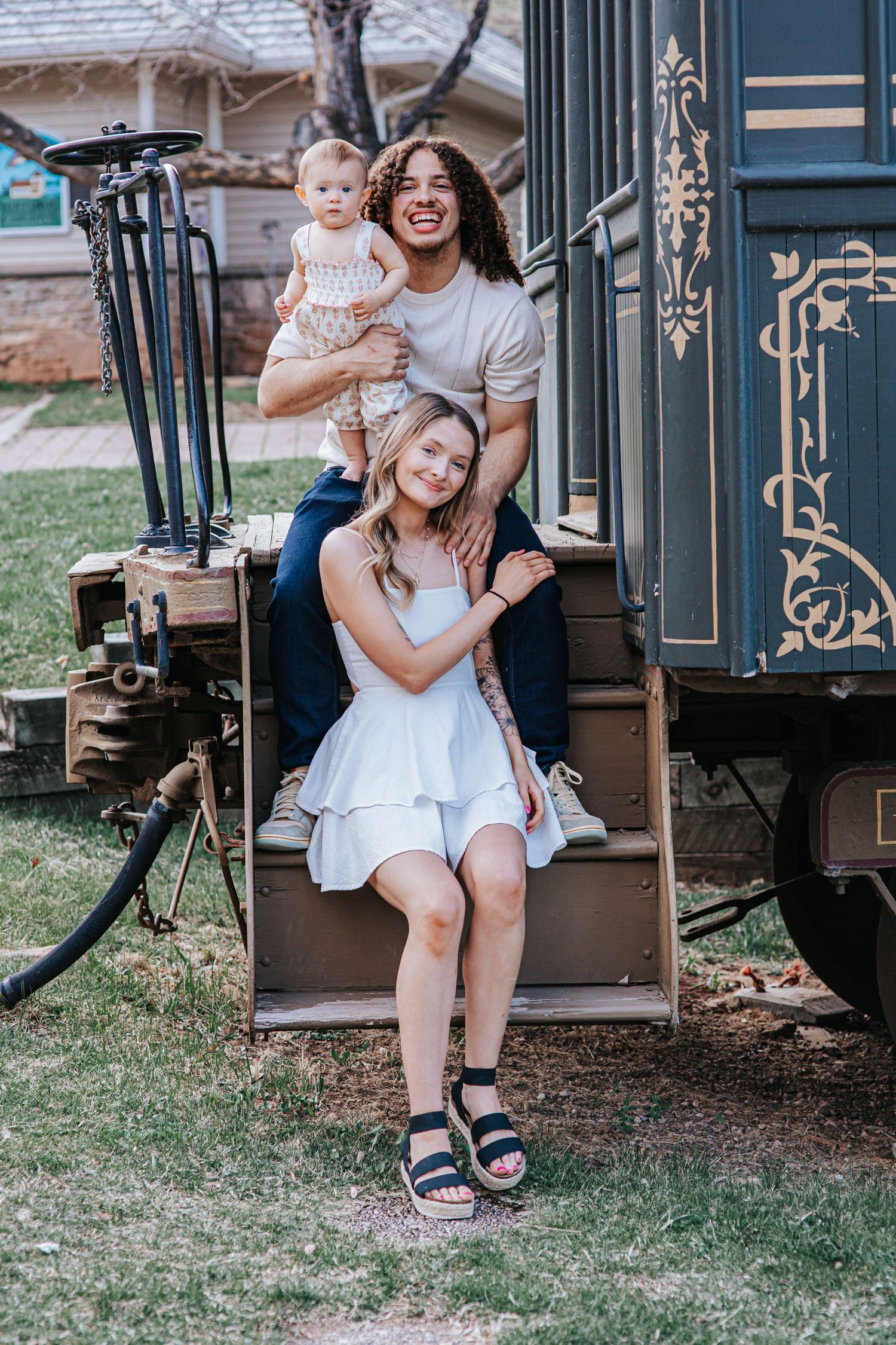 Happy family posing on vintage train steps outdoors, man holding baby, woman in white dress smiling in Spearfish SD