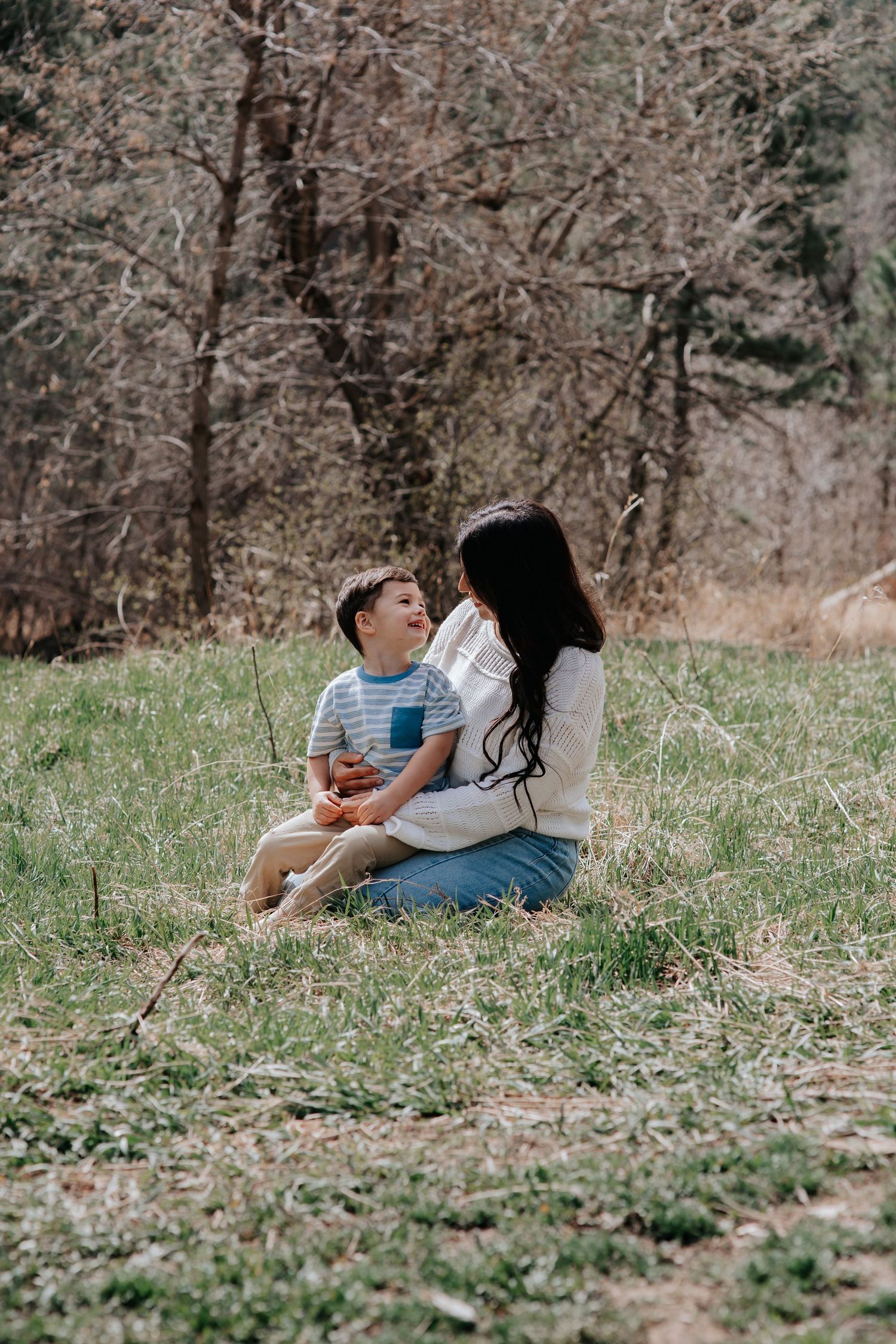 A woman and young boy sit together on green grass in a forest clearing, smiling at each other in a candid outdoor moment.