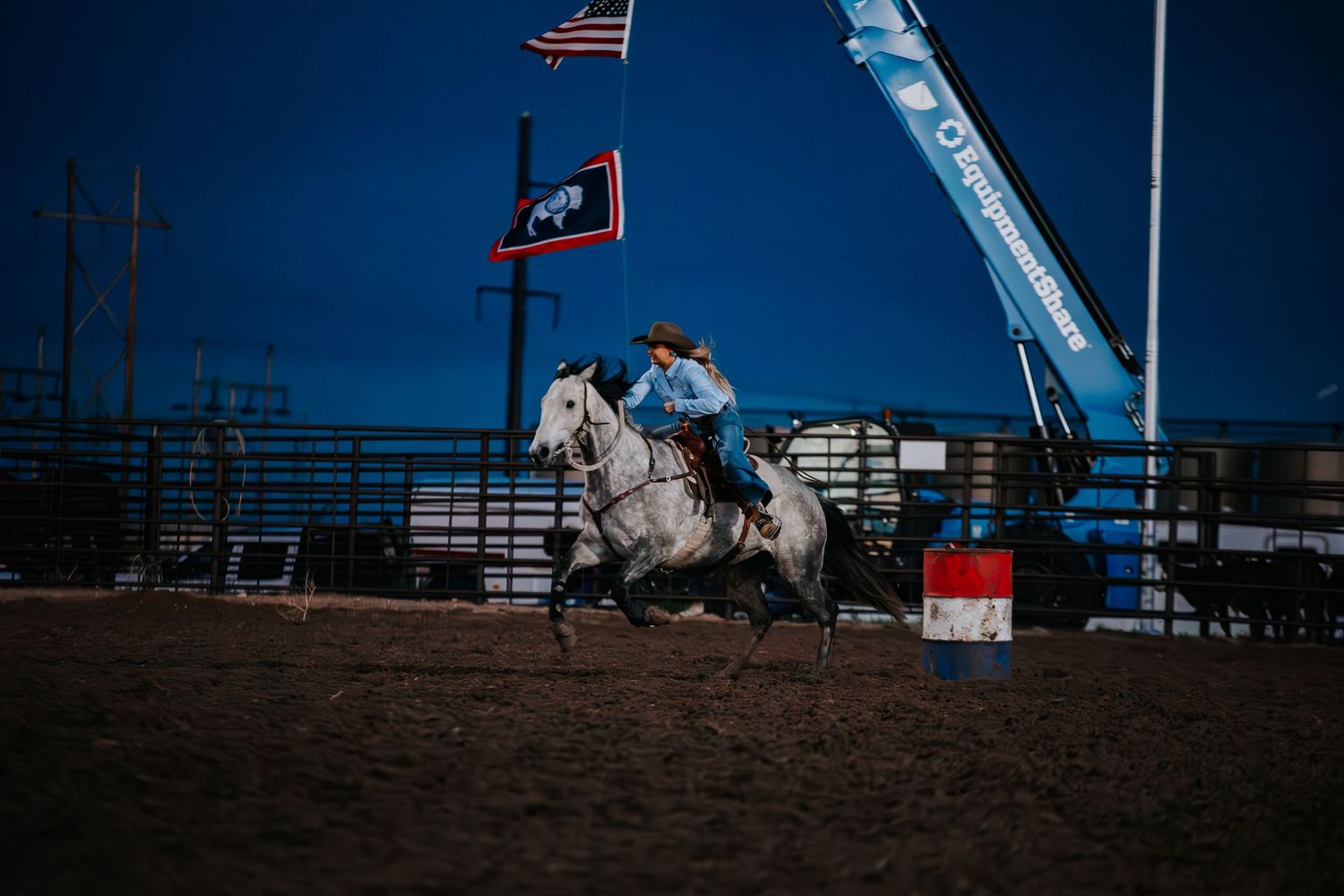 American and state flags wave above a rodeo arena with white fencing under a deep blue sky.