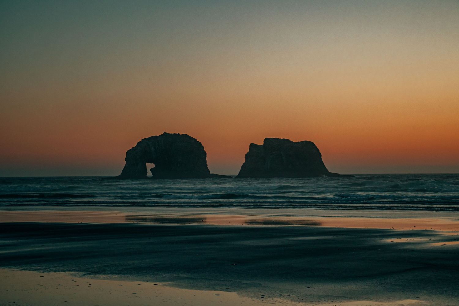 Twin Rocks formation silhouetted against vibrant orange sunset sky at Rockaway Beach, Oregon coast.