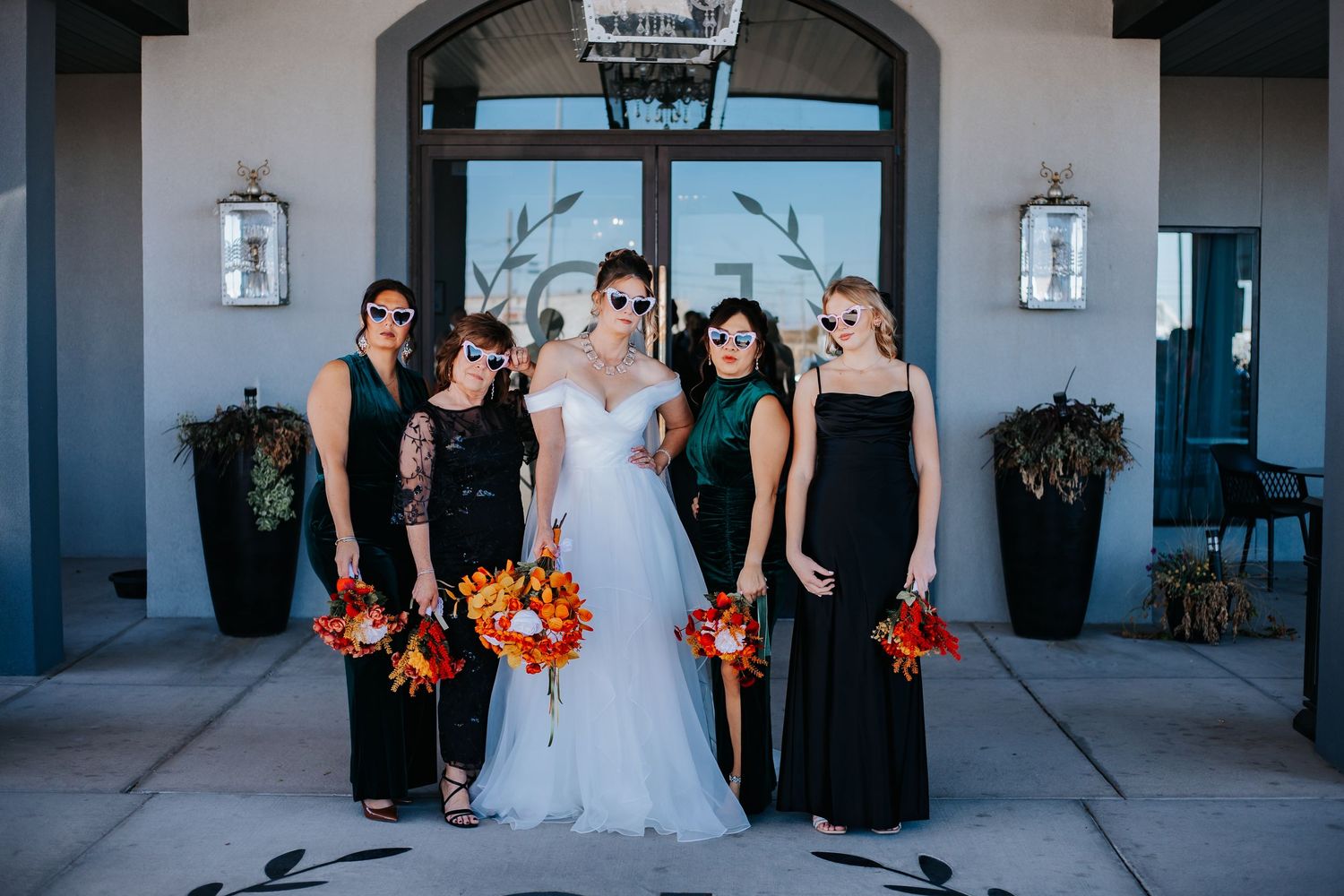 Bridesmaids in black dresses holding orange bouquets pose with bride in white gown at elegant venue entrance.