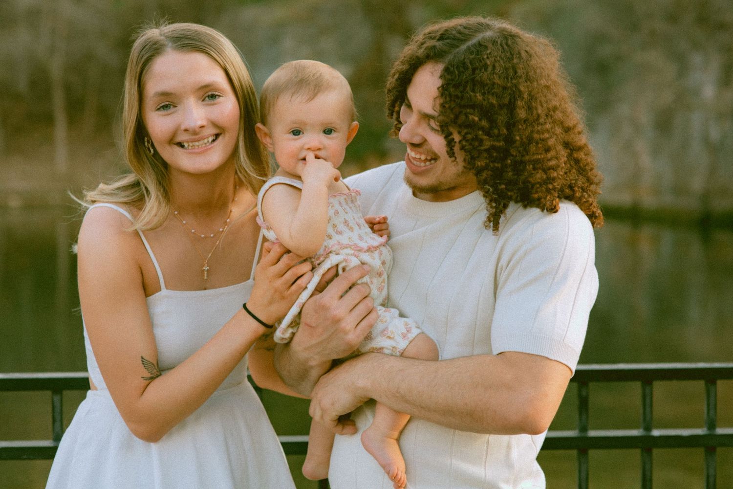 Happy young couple smiling and holding their baby outdoors near a pond in warm golden light.