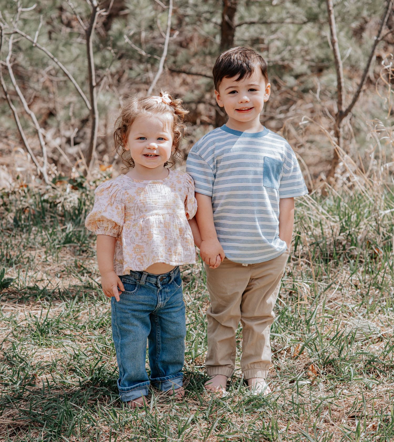 Two young siblings stand together smiling outdoors, girl in floral top and boy in striped shirt.
