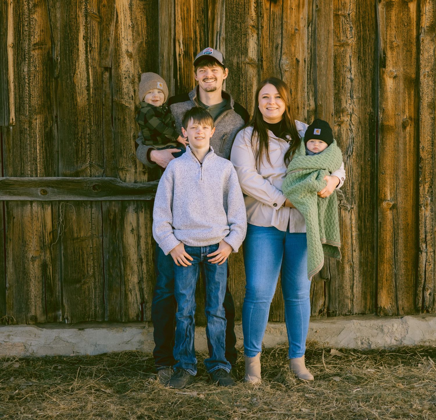 Family portrait session against weathered wooden barn backdrop in coordinated denim and neutral colors in Kimball Nebraska.