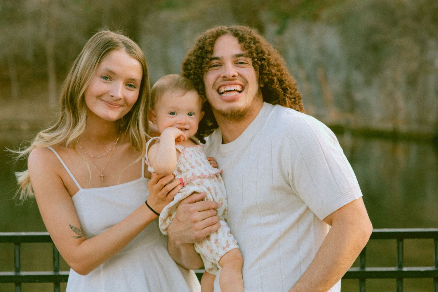 Young couple smiling and holding their baby outdoors near a pond, dressed in white for a family portrait.