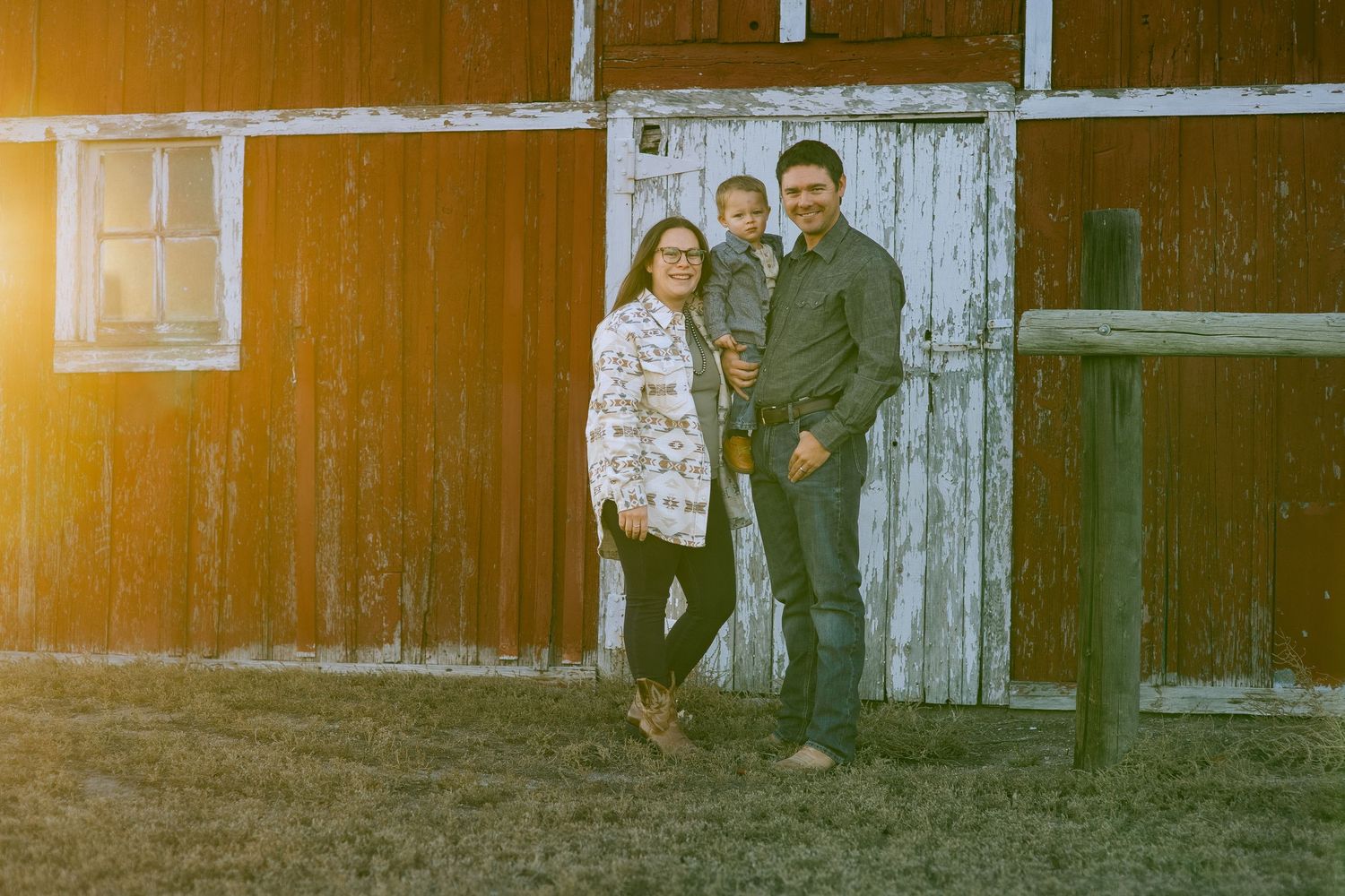 A couple stands together in front of a rustic wooden barn wall on a sunny day in Nebraska for family pictures