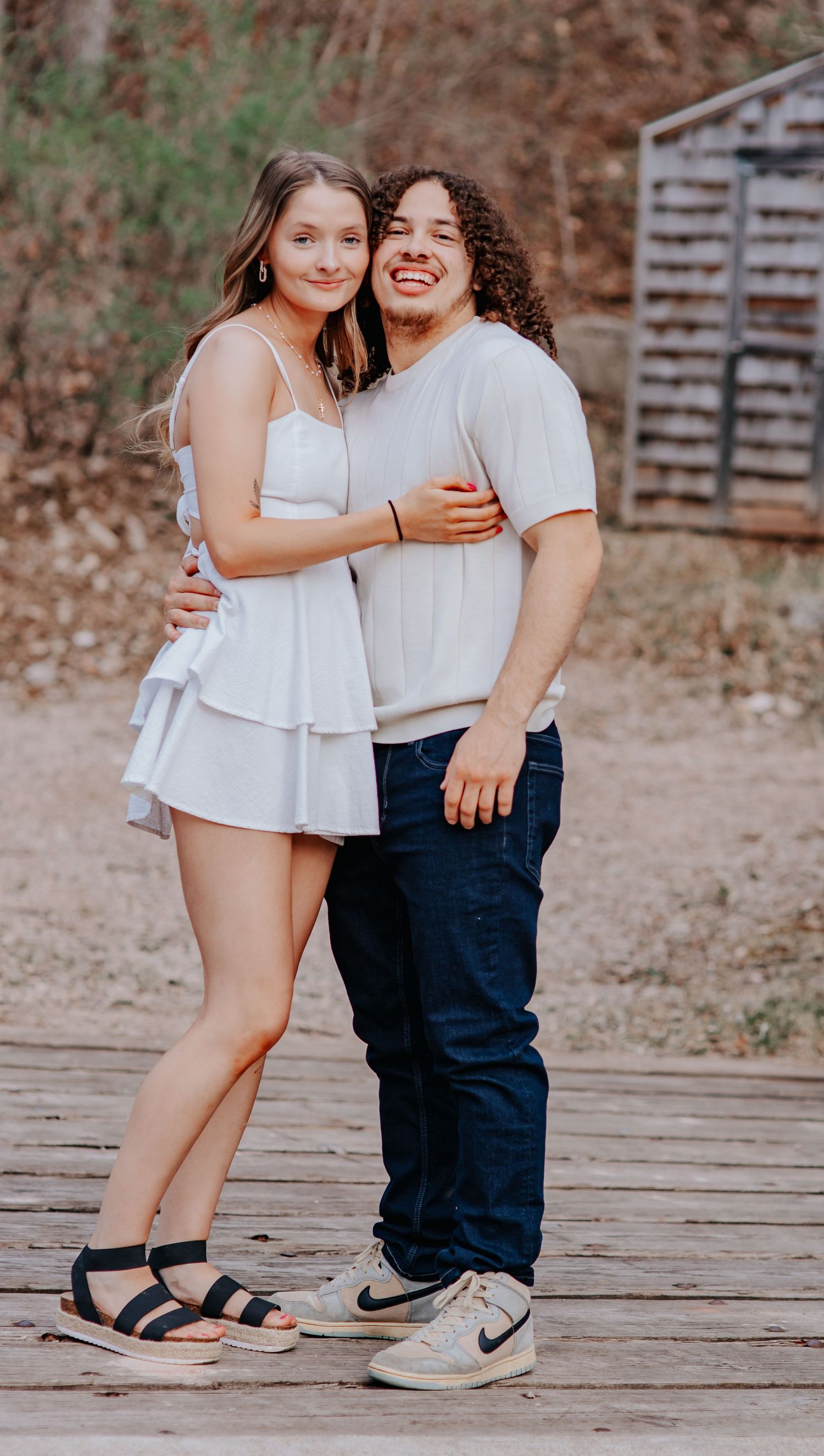 Young couple embracing and smiling on a wooden deck outdoors, woman in white dress, man in jeans and white shirt.