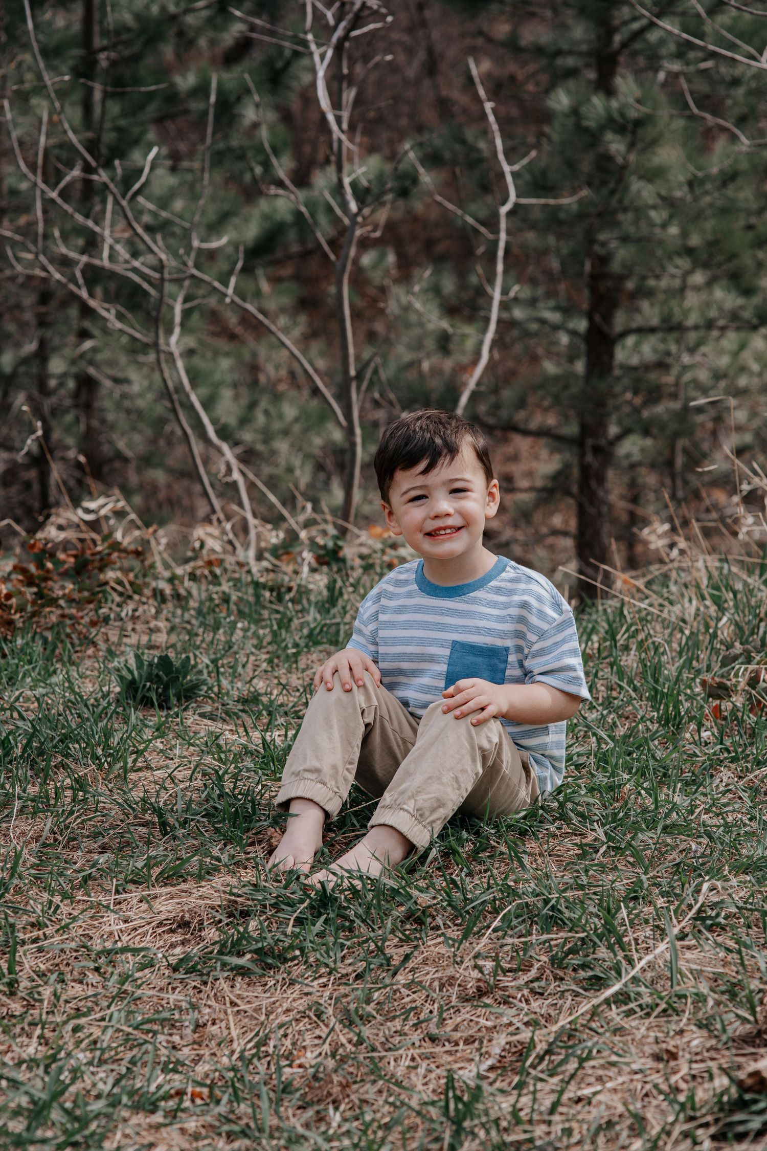 A smiling young boy sits on grass in a forest wearing a striped shirt and khaki pants.