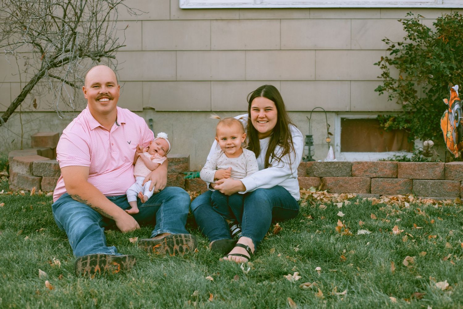 Family sits together on grass with dandelions blooming in their yard during springtime for a lifestyle family session in western nebraska