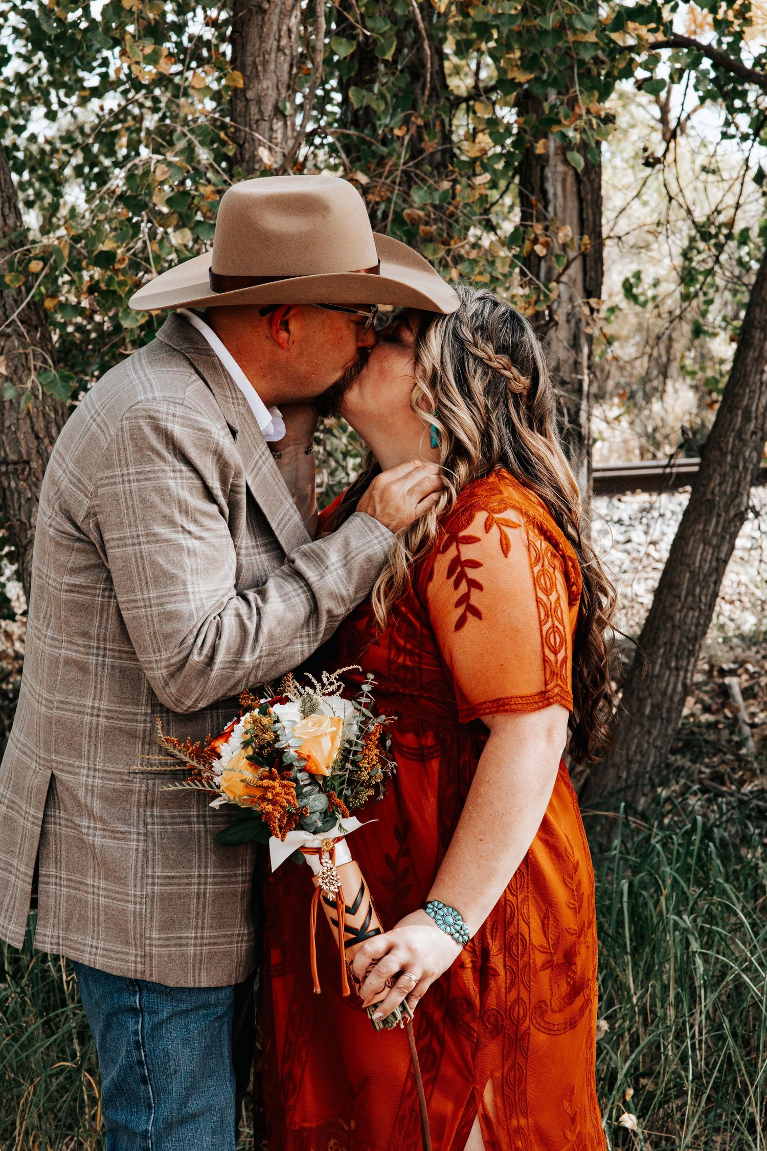 A bride and groom share a romantic kiss outdoors while holding an autumn-themed bouquet with orange and white flowers in Sterling Colorado.