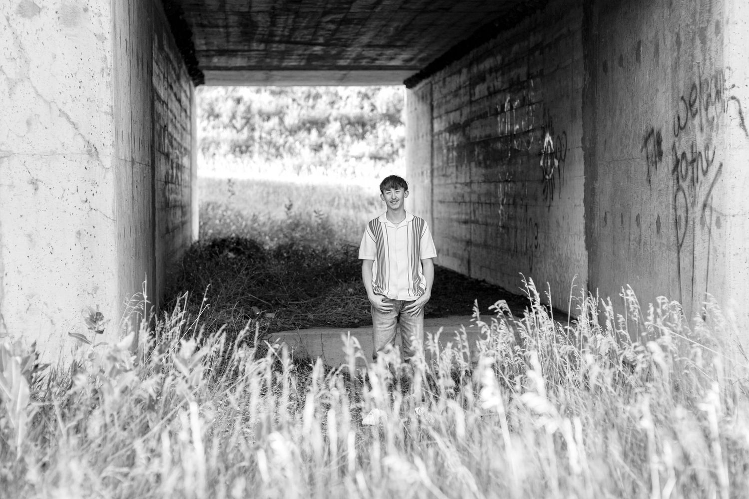 Black and white photo of a high school senior standing in a tunnel surrounded by tall grass and graffiti-covered walls in Nebraska