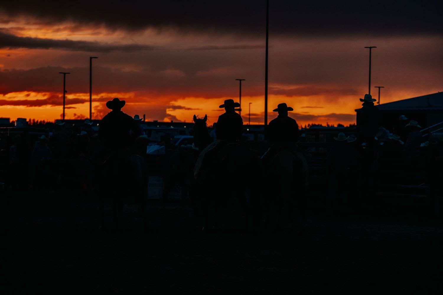 Dramatic orange sunset silhouettes power lines and buildings against a vibrant sky at a rodeo venue.