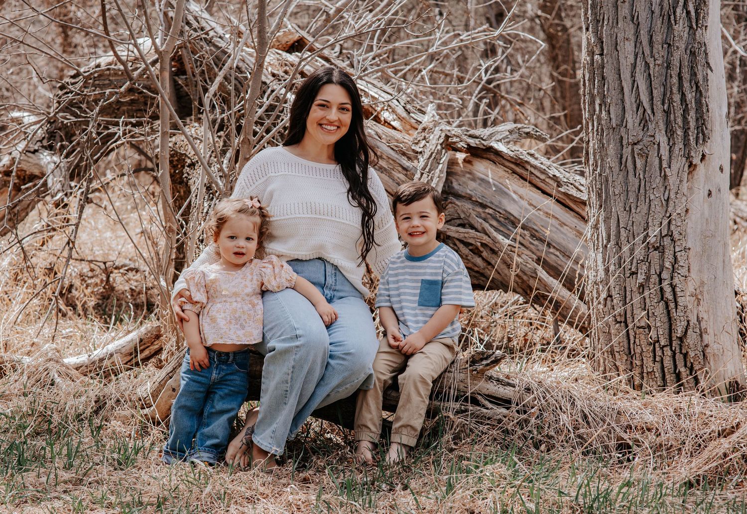 A mother sits outdoors with her two young children against a rustic fallen tree in a natural woodland setting.