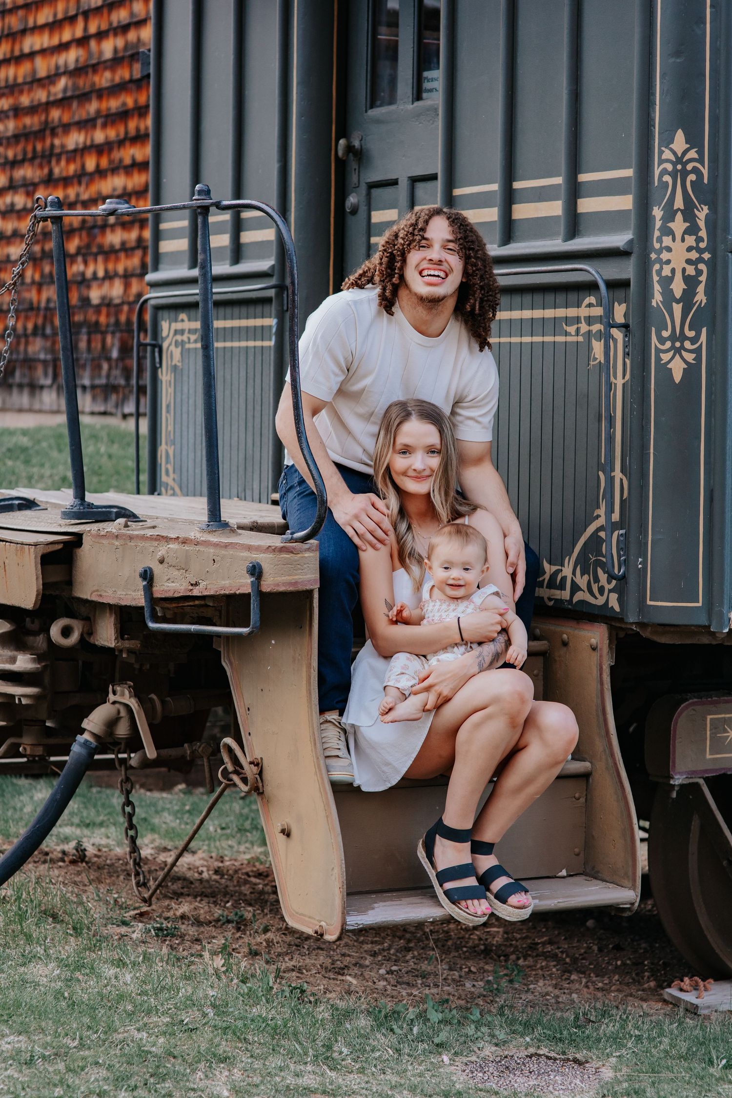 Young family with curly-haired man, woman, and baby sitting on vintage train car steps, smiling outdoors.