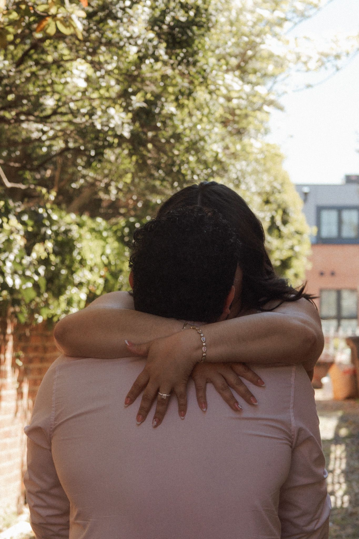 Two people sharing a warm embrace outdoors, with lush green trees and a brick building in the background.