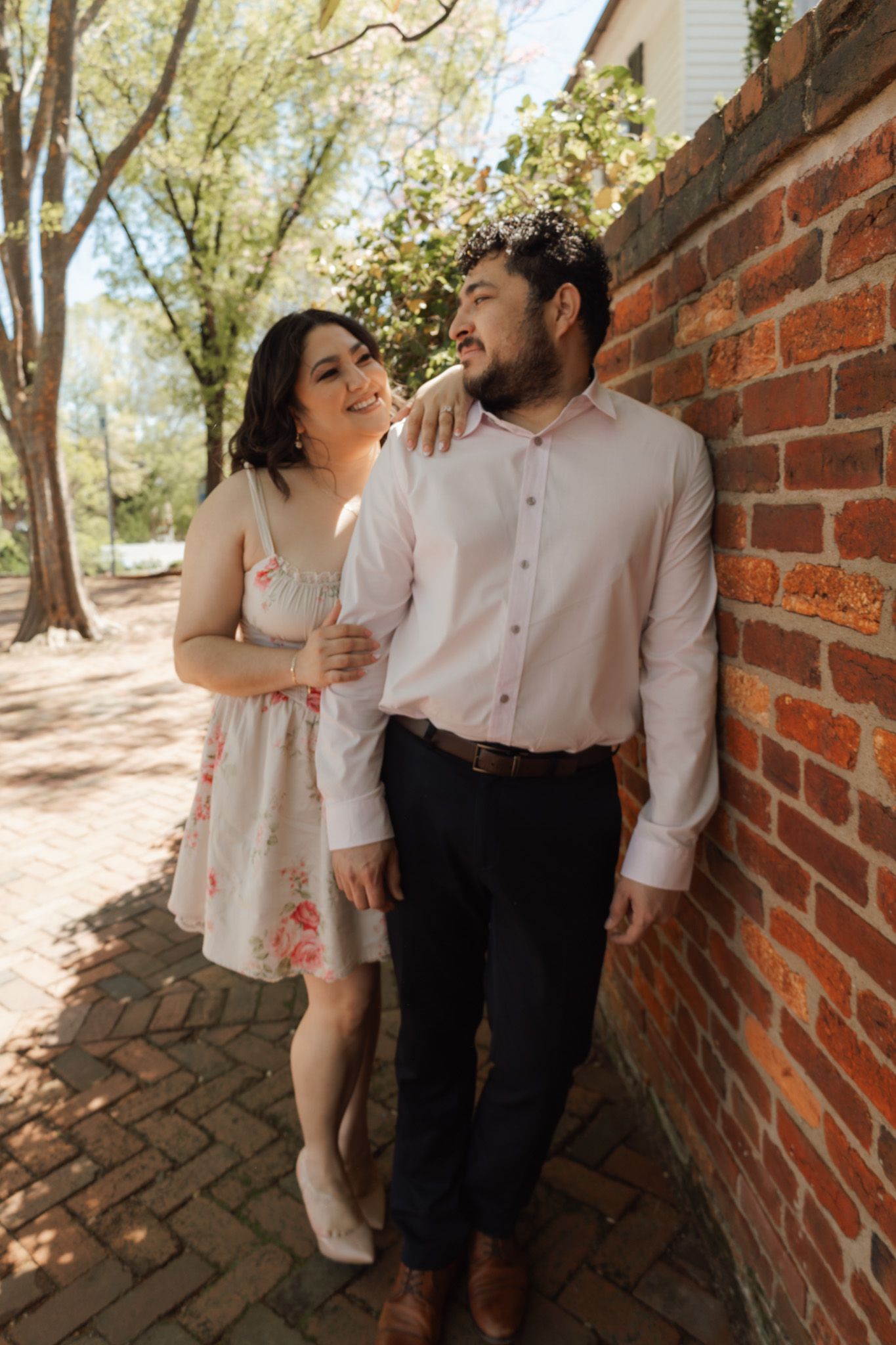Couple posing by a brick wall outdoors, woman in floral dress embracing man in pink shirt on a sunny day.
