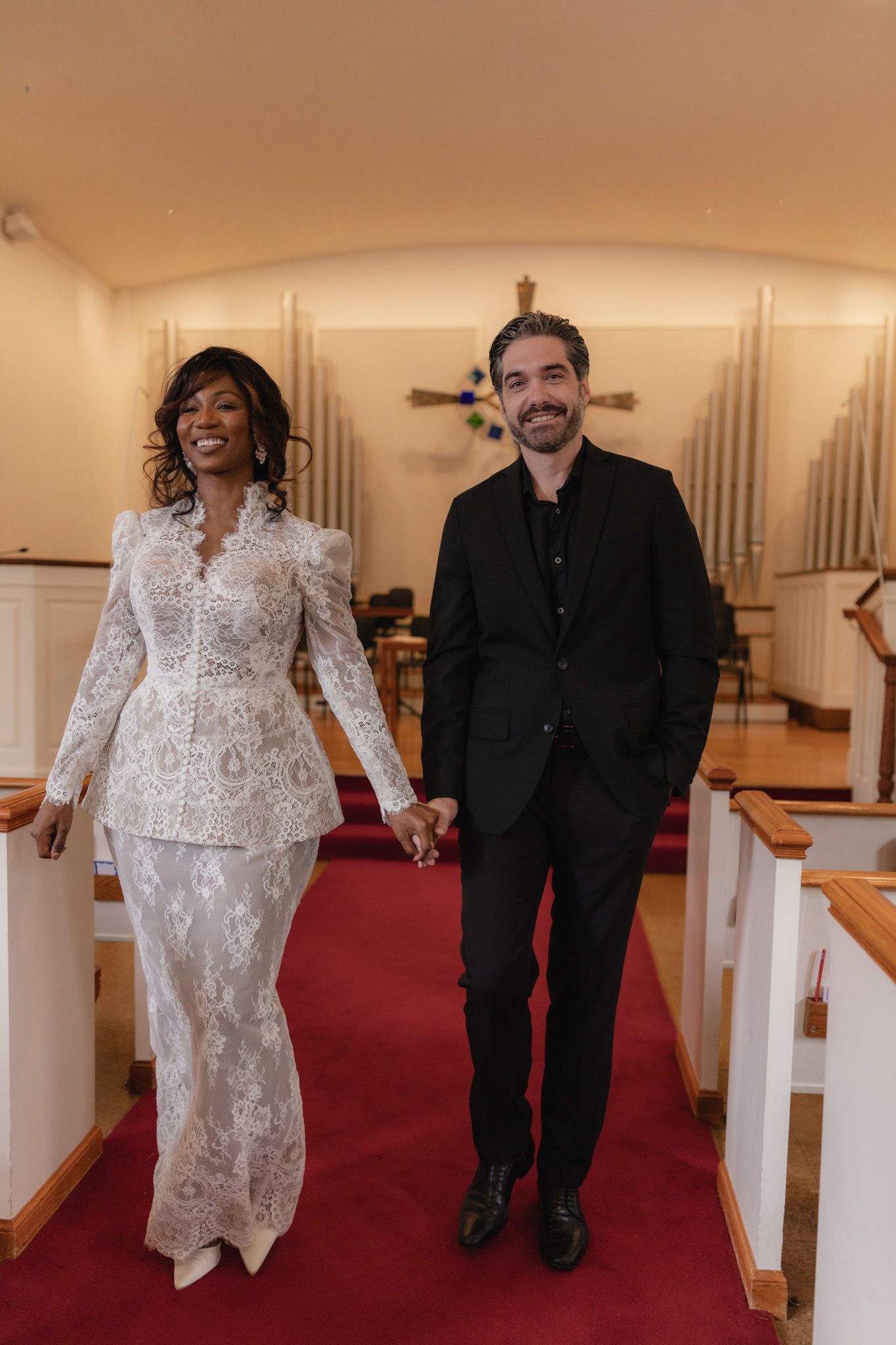 Interracial couple holding hands walking down church aisle, bride in white lace suit, groom in black suit.
