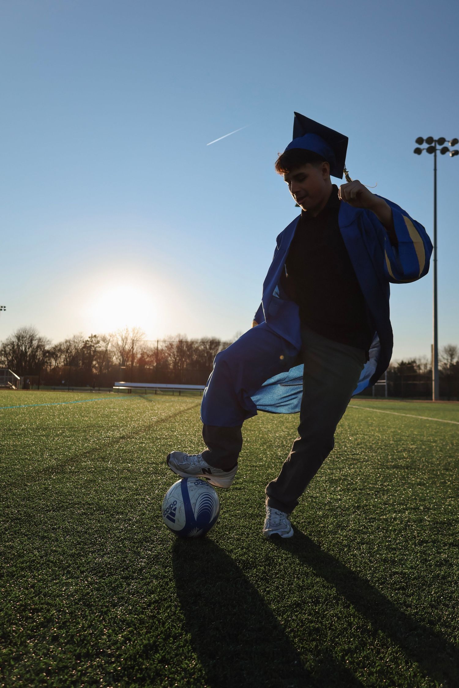 Graduate in blue cap and gown kicking soccer ball on field at sunset.