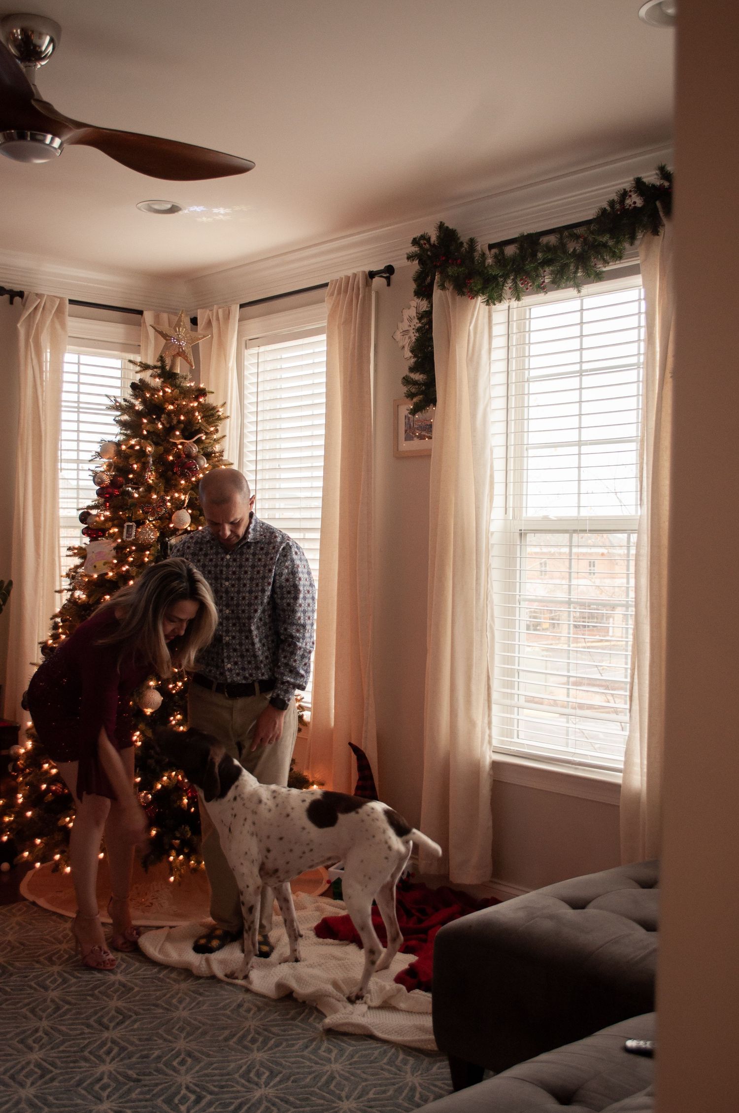Couple and spotted dog by decorated Christmas tree in cozy living room with garland and warm holiday lights.