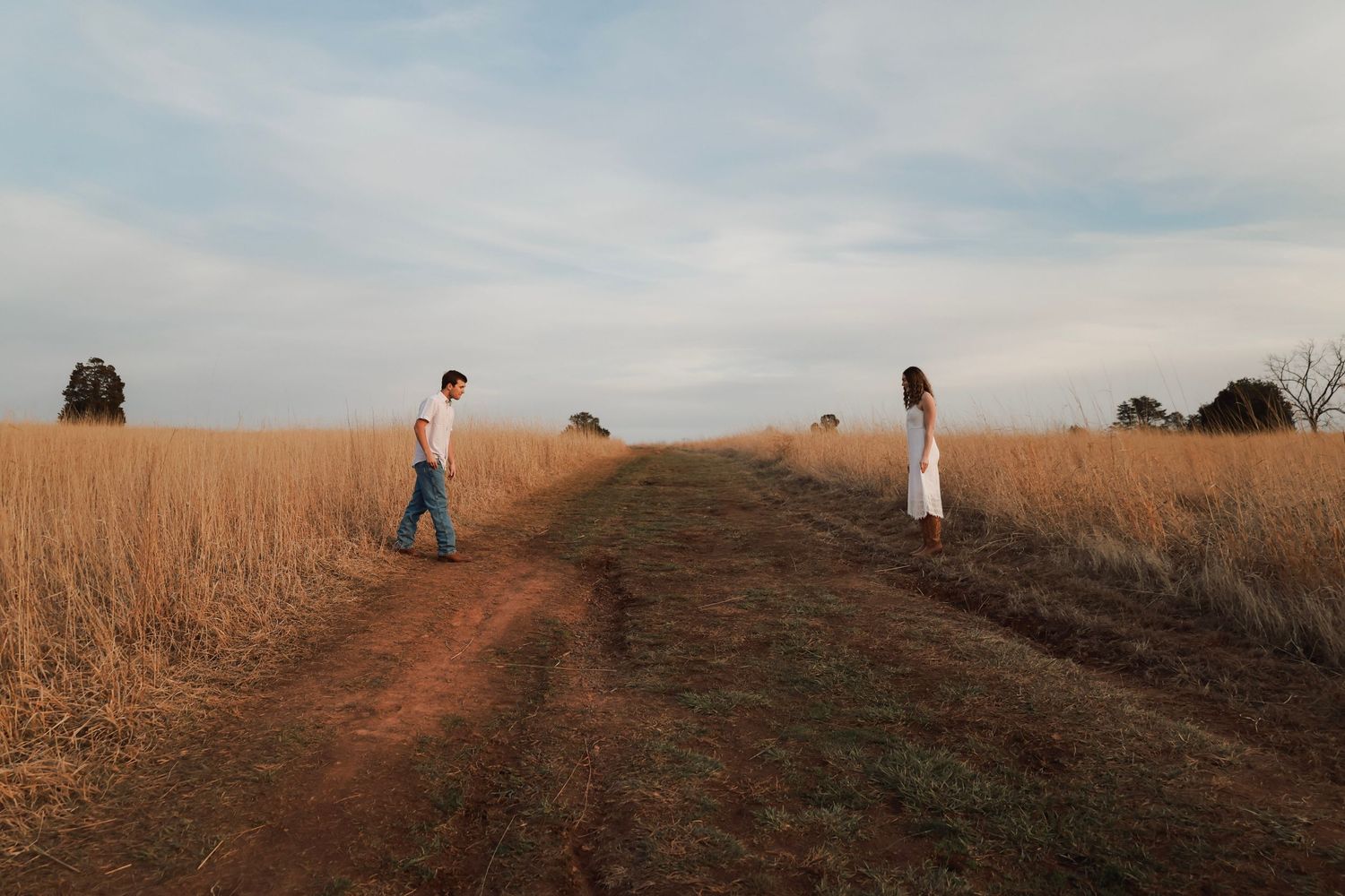 A man and woman stand apart on a dirt path through golden grasslands under a cloudy sky.